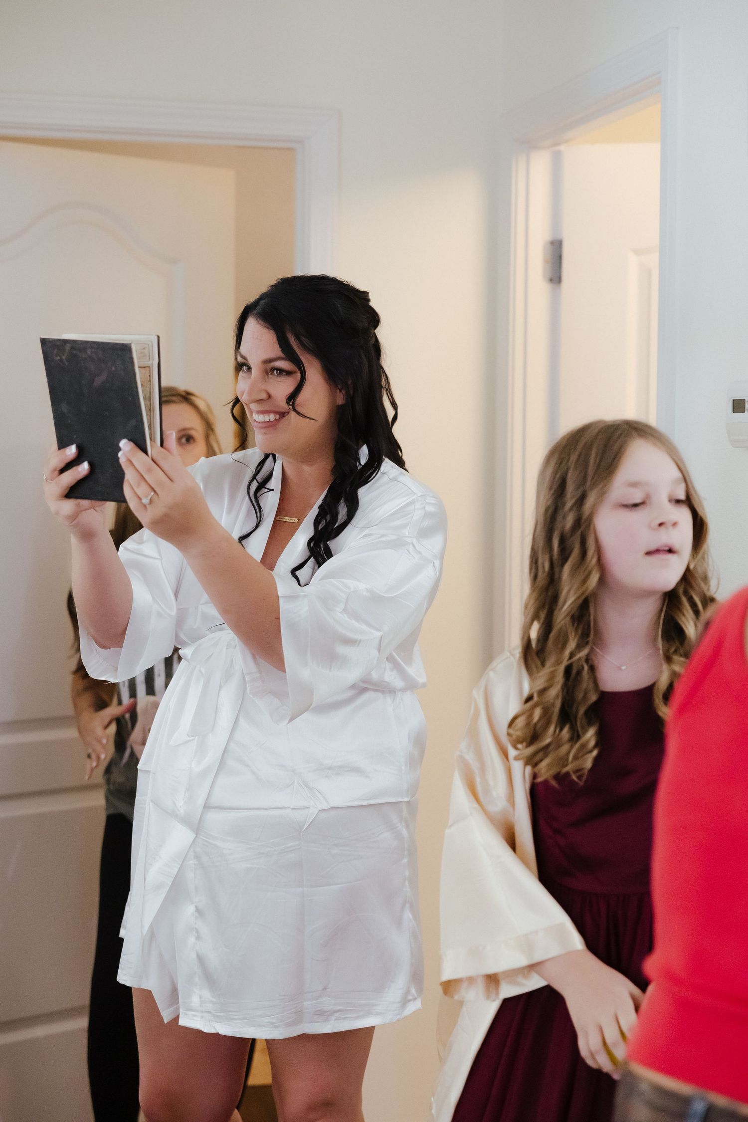 Bride in white robe smiling while holding mirror during wedding morning preparations with bridesmaids.
