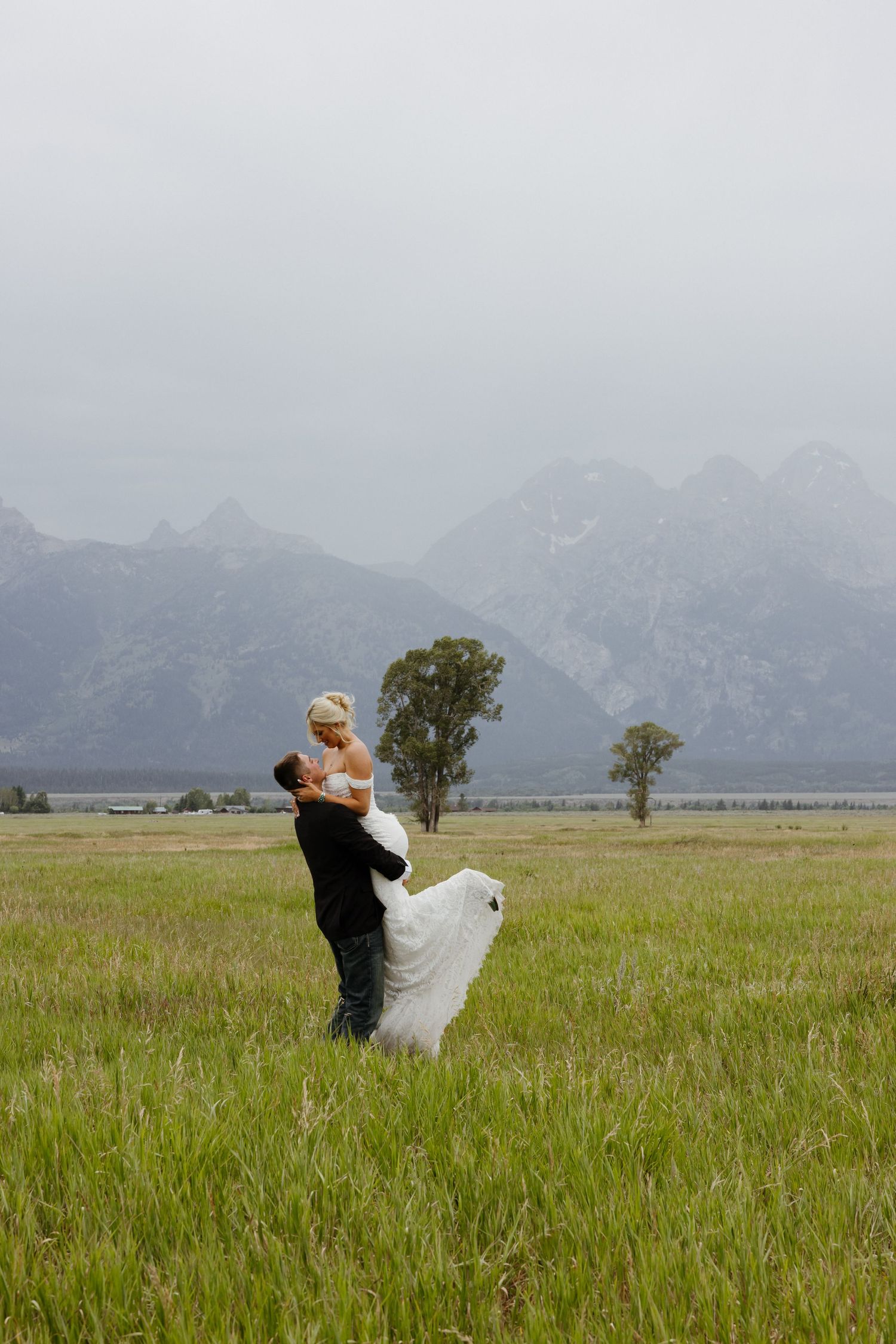 Groom lifts bride in white gown in green meadow with misty Grand Teton mountains backdrop.