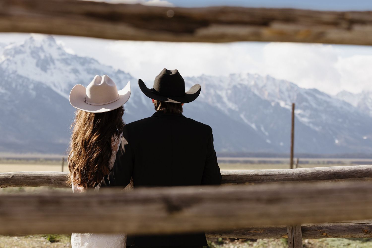 Couple in cowboy hats sitting on a wooden fence with snow-capped Grand Teton mountains in the background.