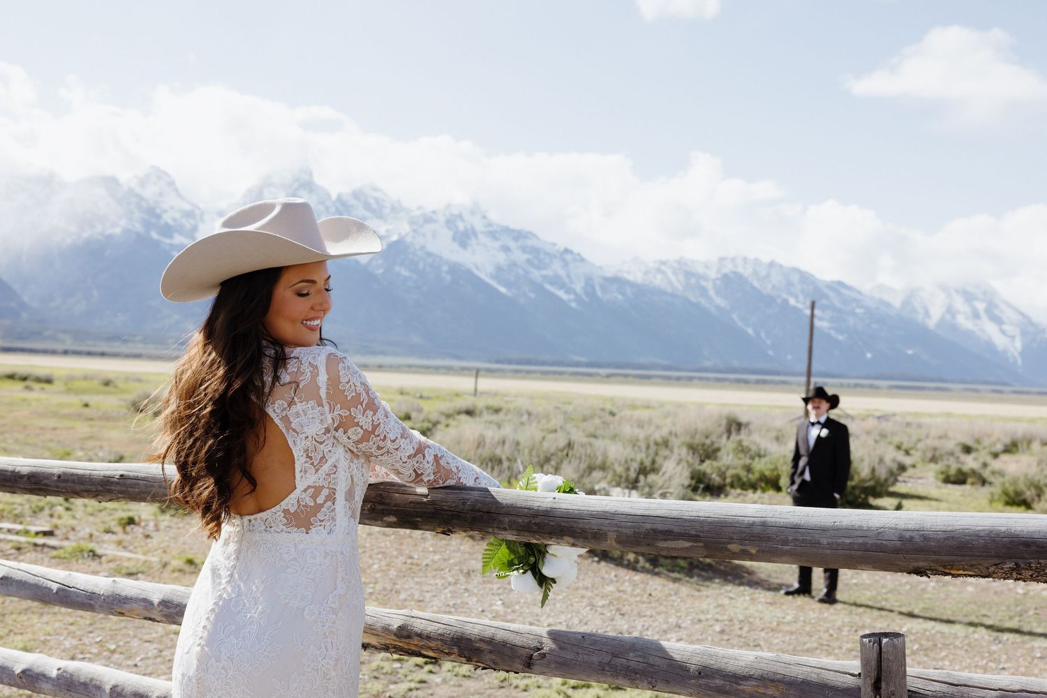 Western bride in lace dress and cowboy hat leans on wooden fence with Grand Teton mountains in background.