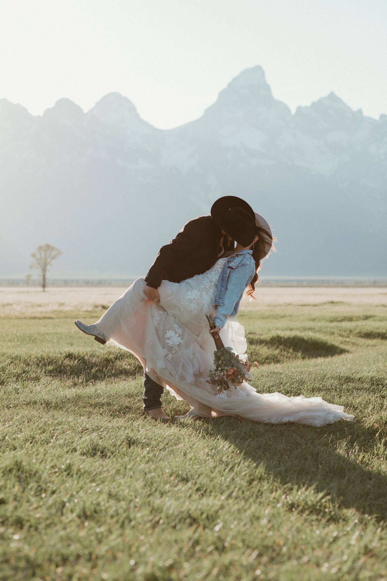 Groom dips bride in white lace wedding gown on green meadow with Grand Teton mountains behind.