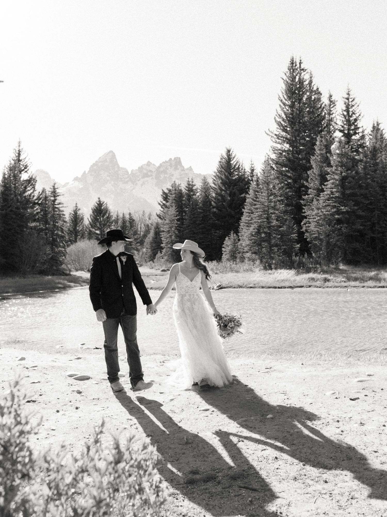 Black and white photo of bride and groom in cowboy hats holding hands near a river with mountain backdrop.