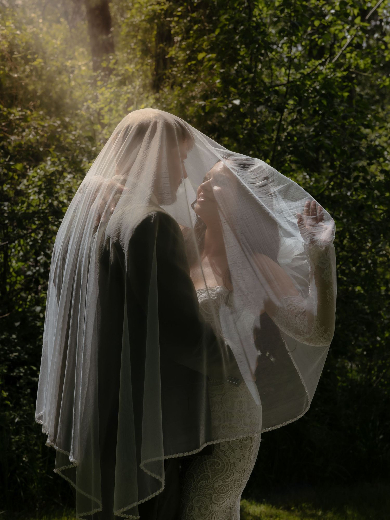 Romantic photo of bride and groom kissing under a flowing veil in golden sunlight among green trees.