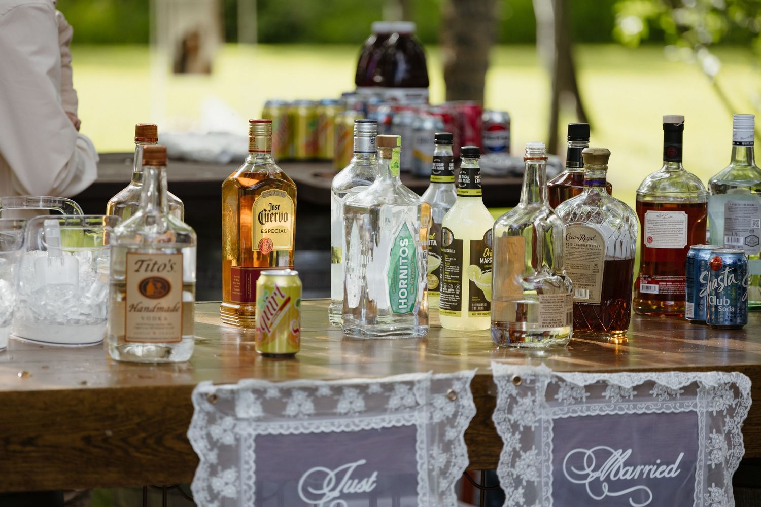 Rustic outdoor wedding bar display featuring assorted liquor bottles with Just Married chalkboard signs on lace tablecloth.