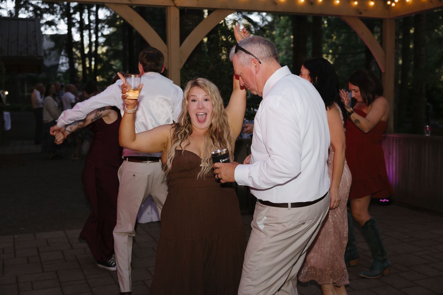 Excited wedding guests laugh and dance together under string lights at an outdoor evening forest reception.