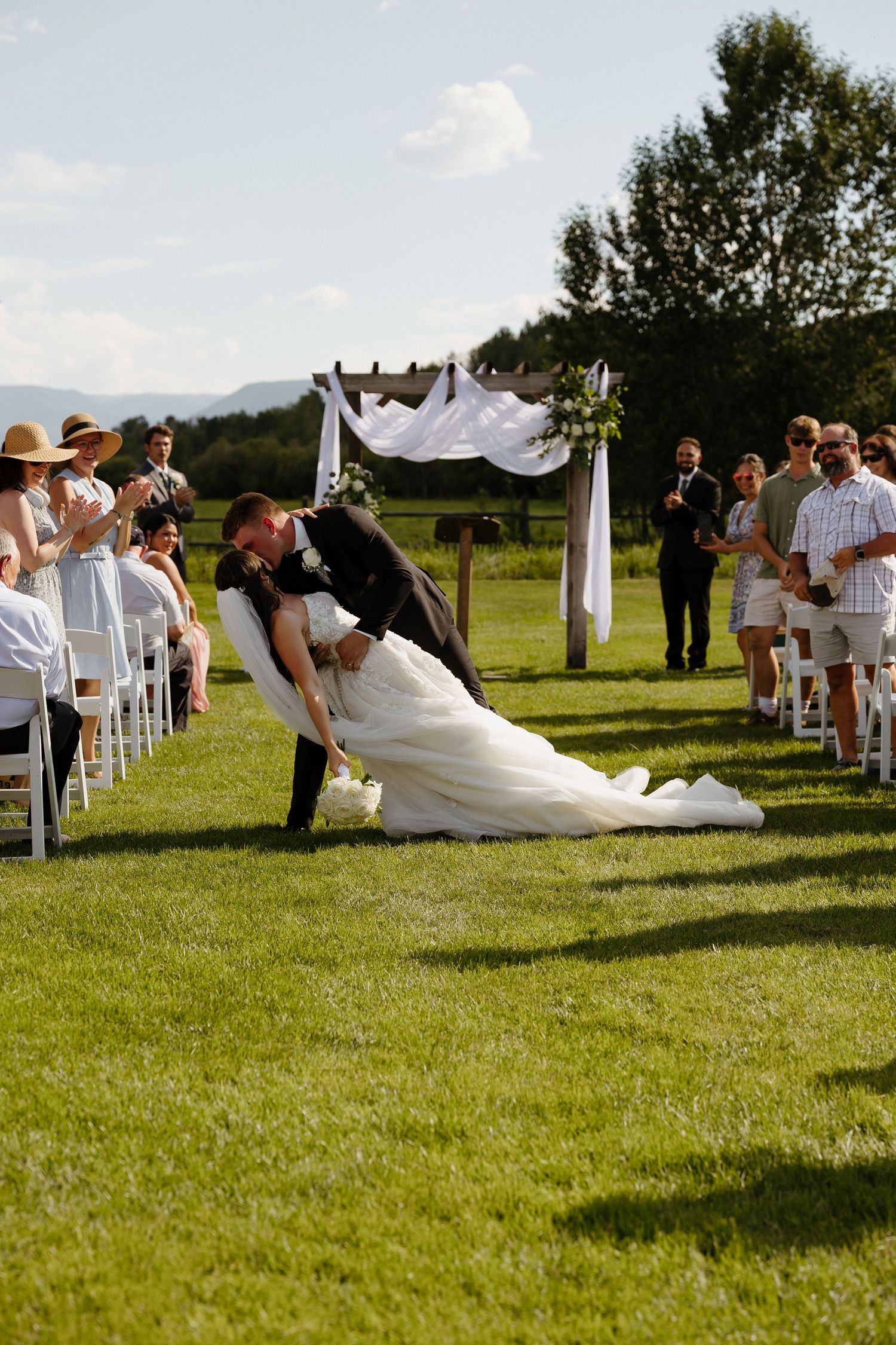 Groom dips bride for romantic kiss in wedding aisle while guests applaud outdoors.