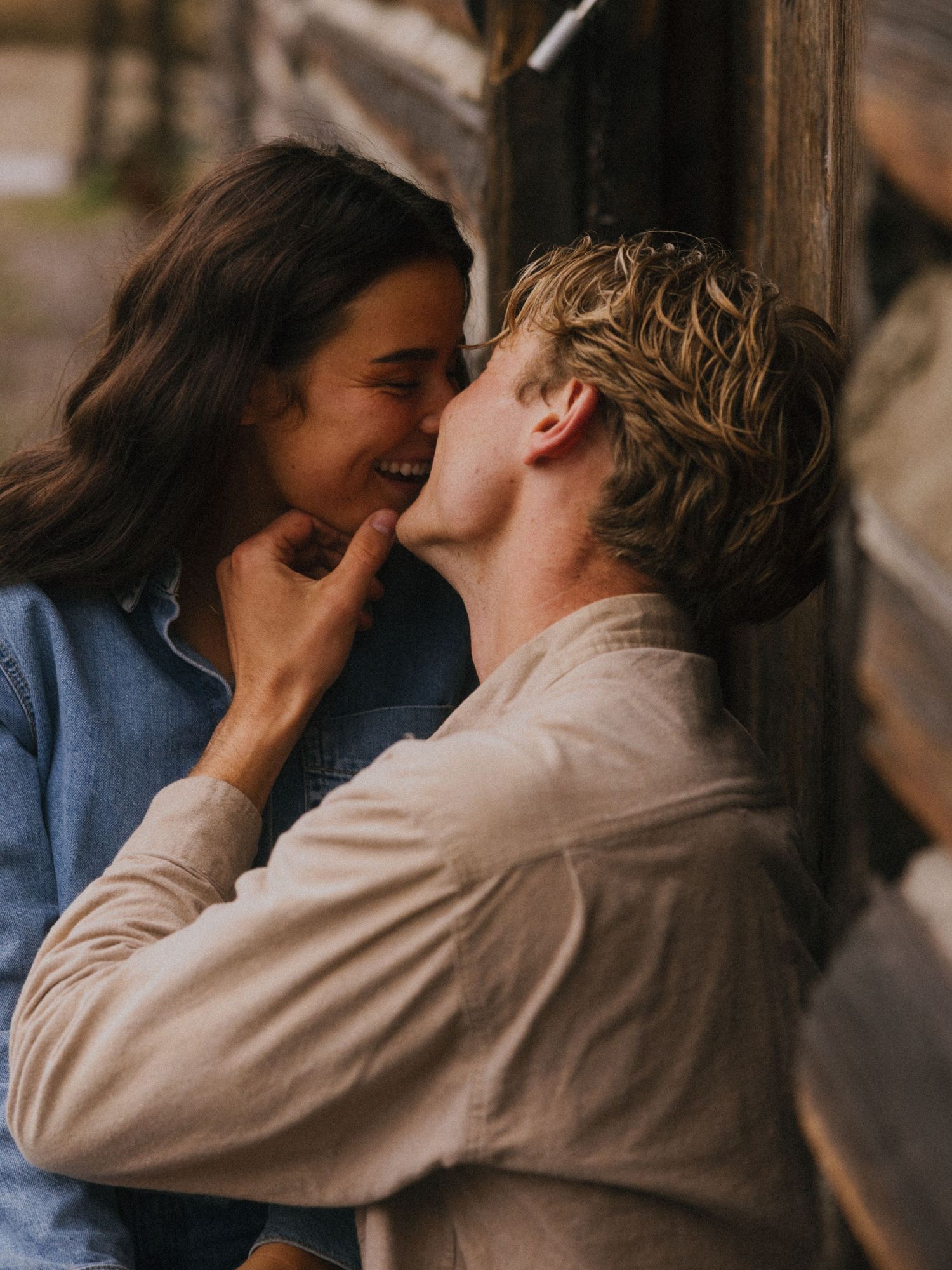 Couple sharing an intimate moment laughing and almost kissing against a rustic wooden wall outdoors.