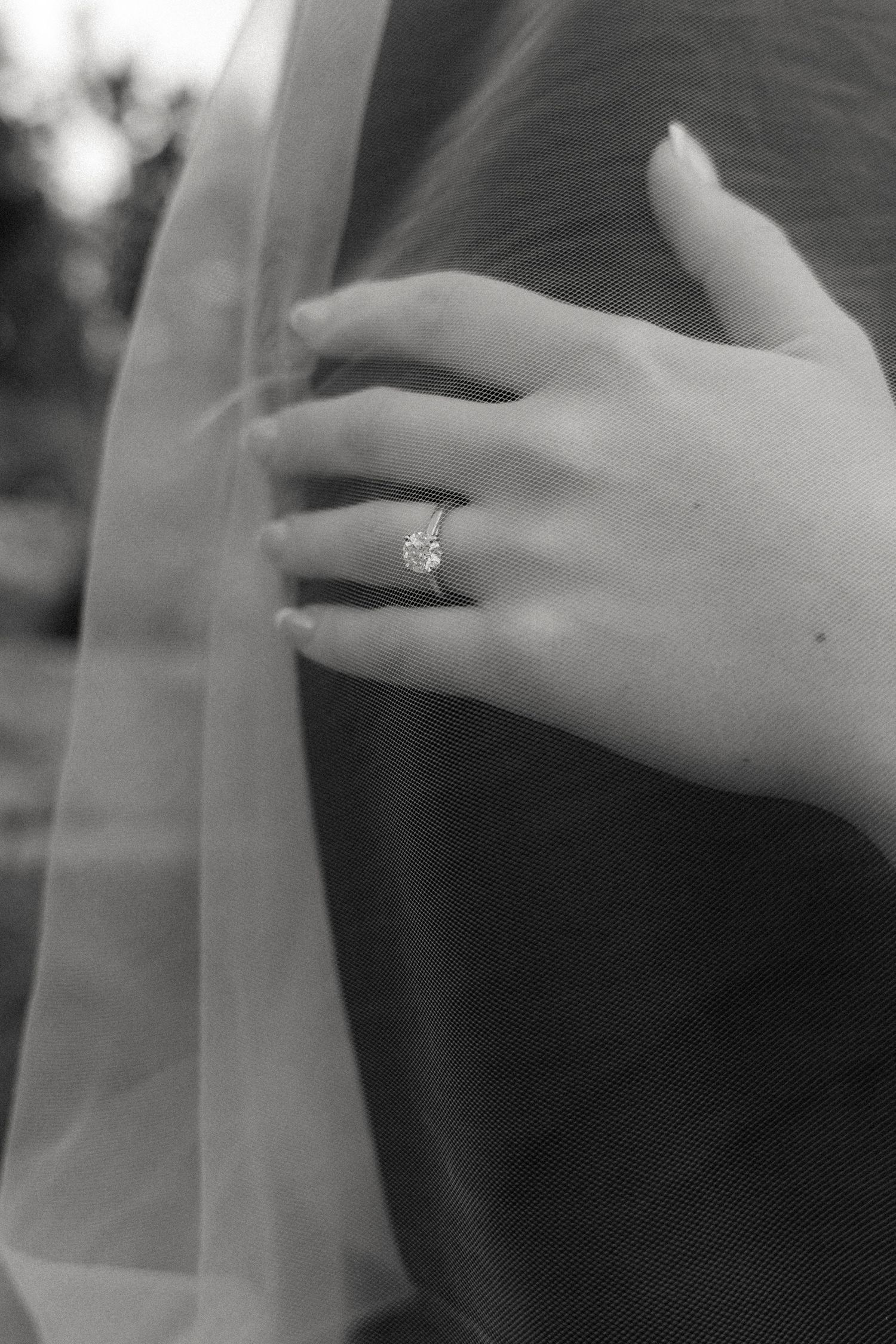 Black and white close-up of bride's hand with solitaire diamond ring resting on groom's chest near veil.