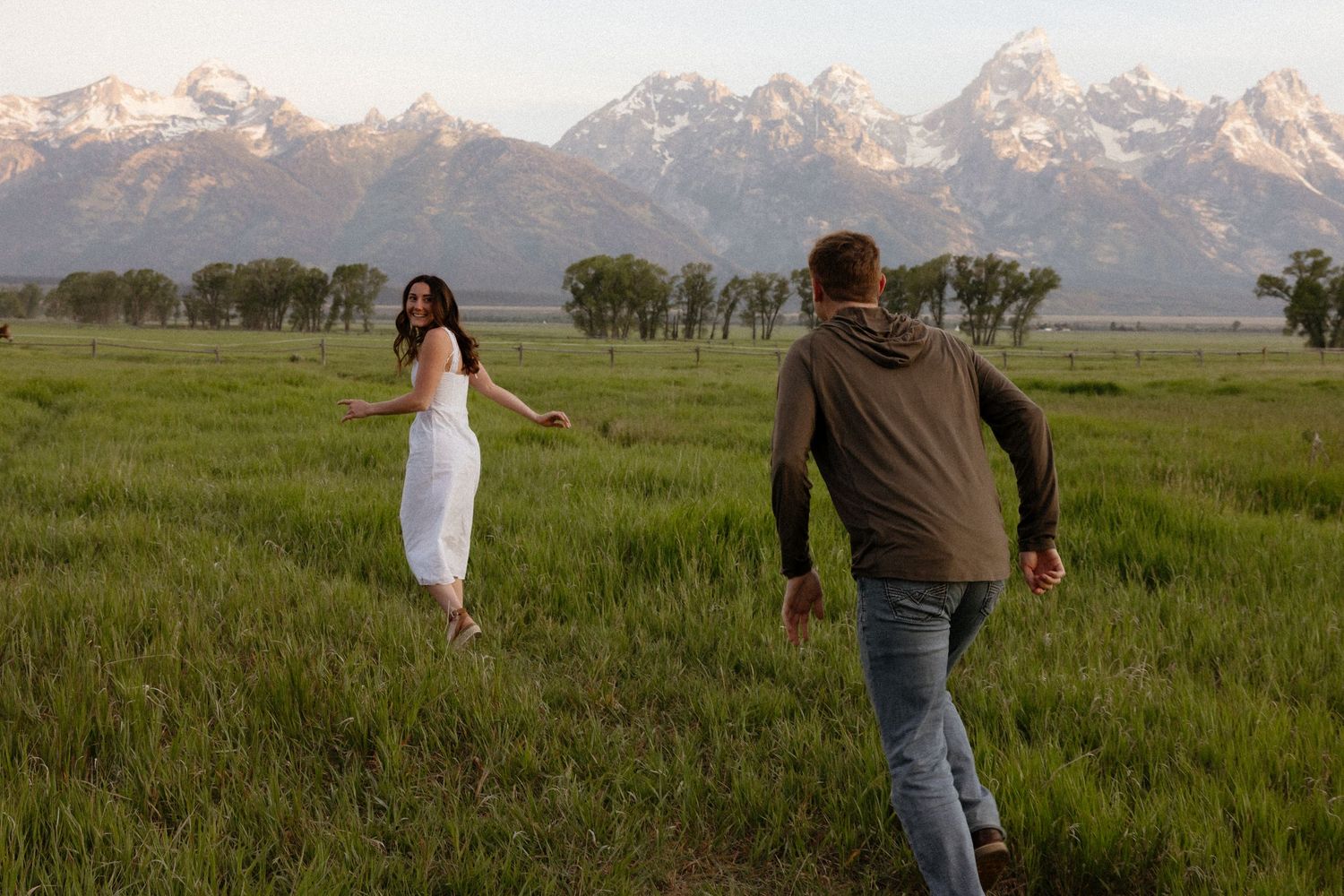 Woman in white dress reaching for her partner's hand in a green field with Teton mountains.