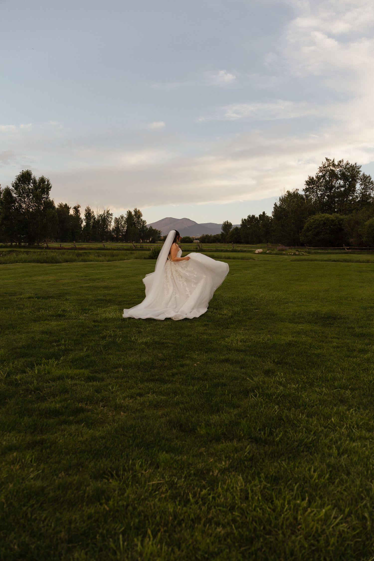 Bride twirling in a flowing white gown and veil on a lush green field at golden hour with mountains in the background.