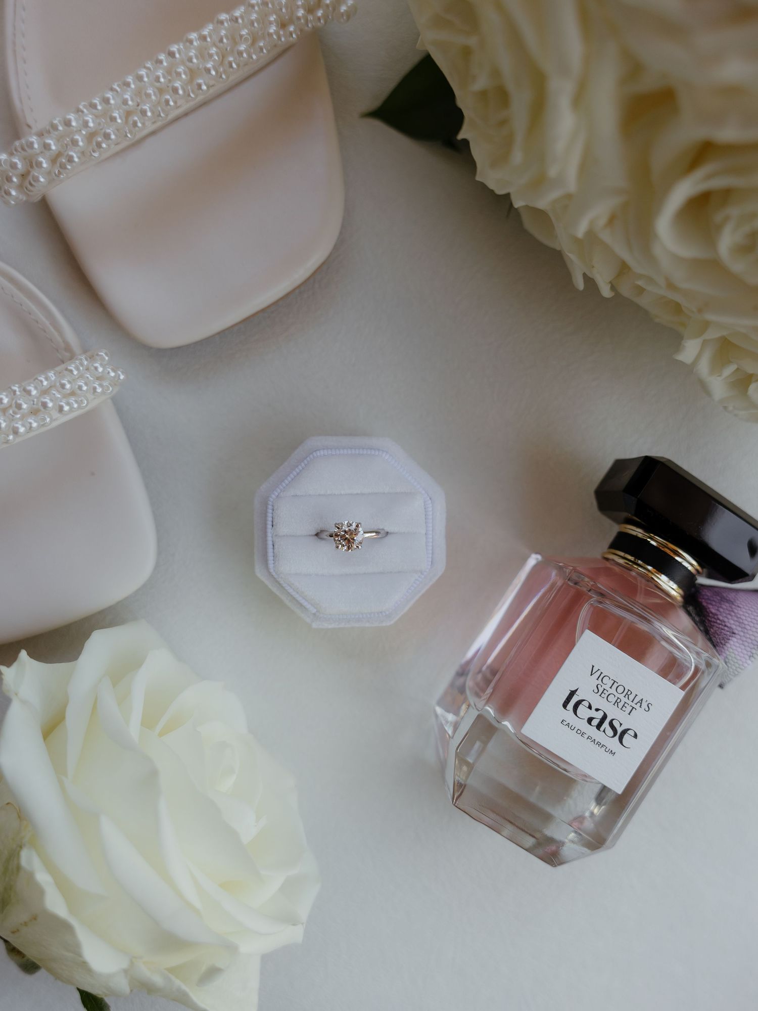 Bridal detail flat lay with rose gold ring in white box, Victoria Secret perfume, and pearl heels.