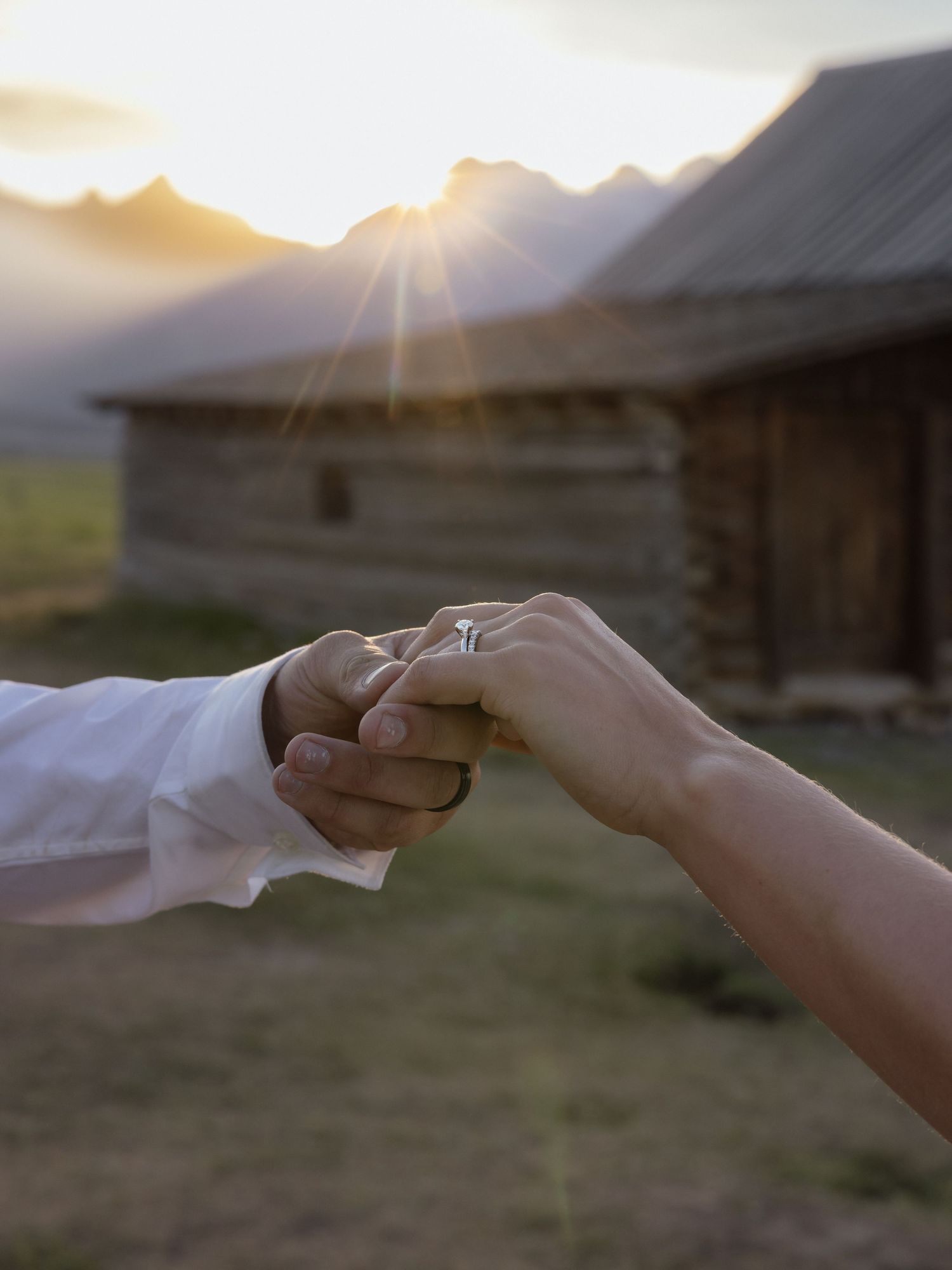 Close-up of bride and groom holding hands with wedding ring visible at golden sunset near cabin.