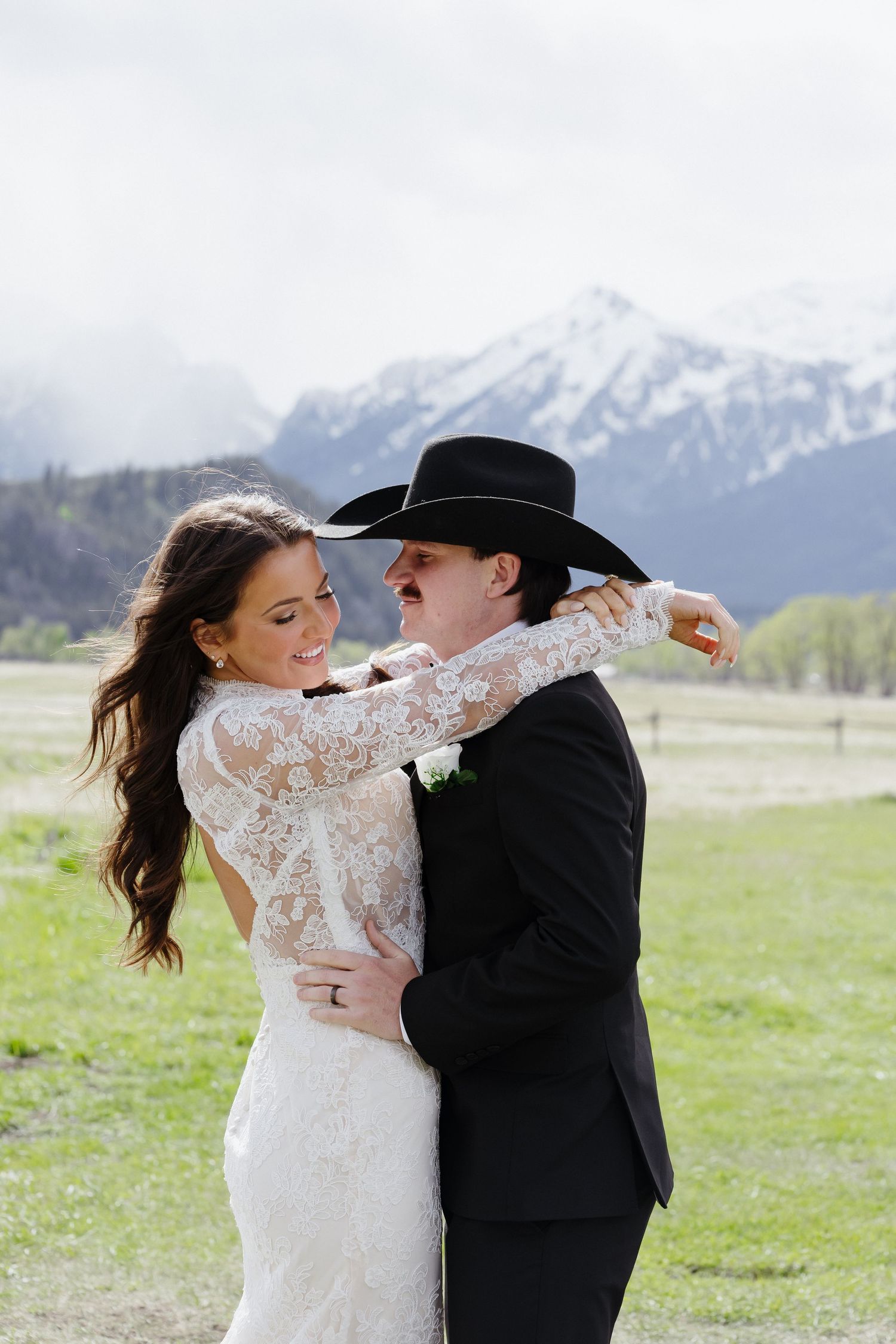 Smiling bride in lace gown embraces groom in black cowboy hat with snowy mountain peaks behind them.