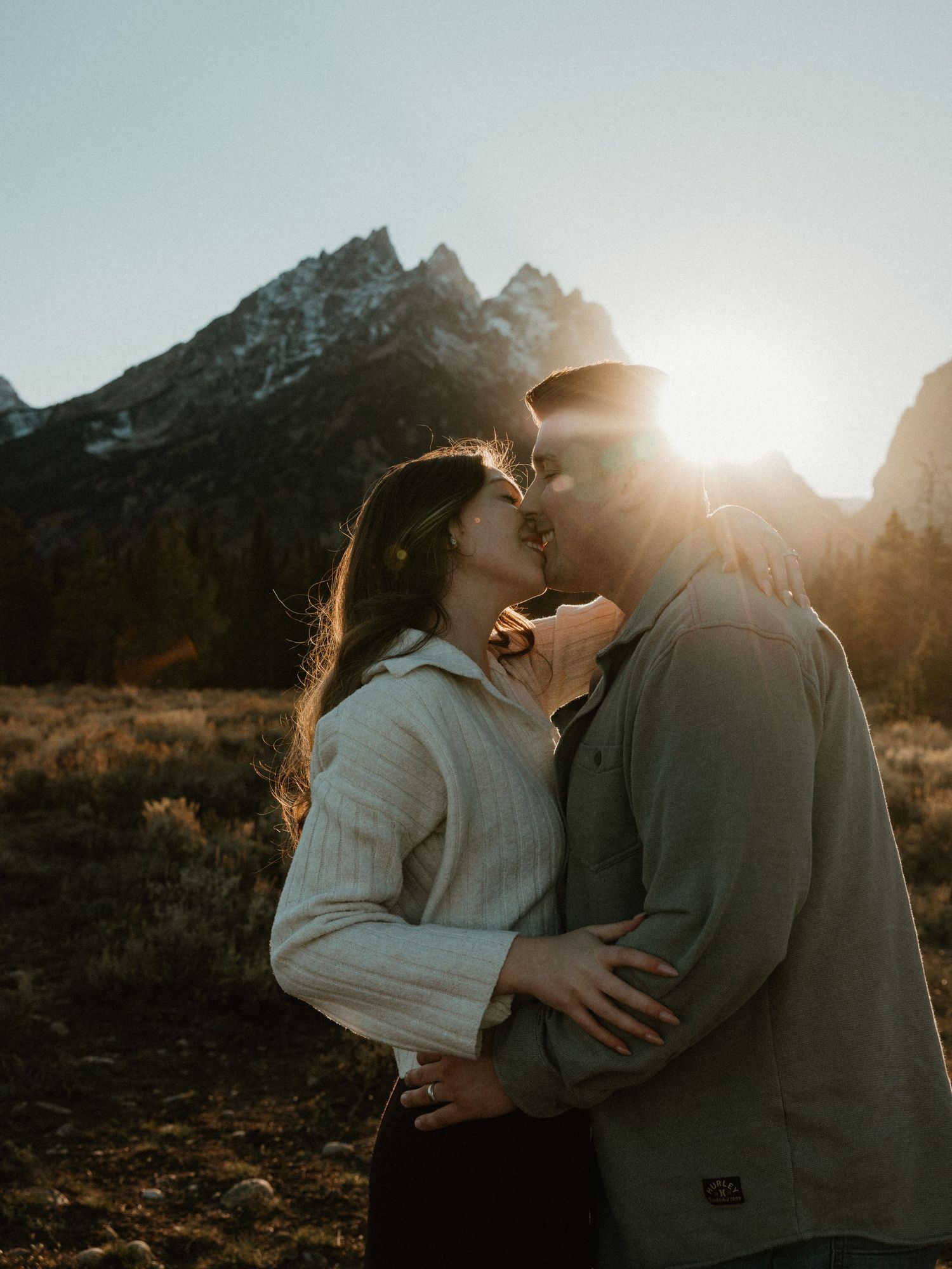 Couple sharing a romantic kiss with golden sunburst and rugged mountain peaks behind them at sunset.