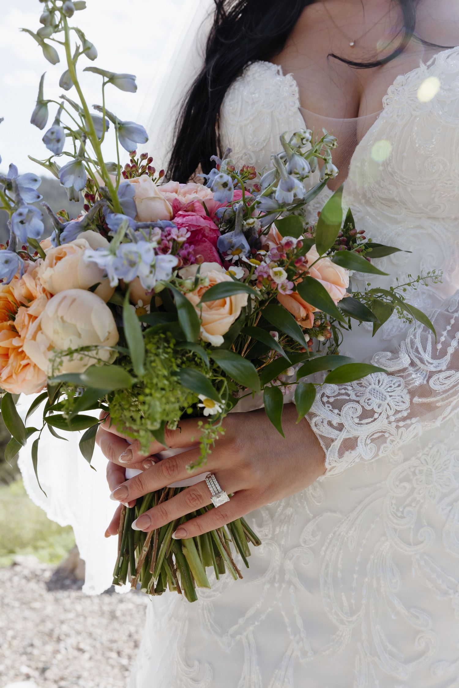Bride in lace wedding gown holding vibrant bouquet of peach roses and blue wildflowers.