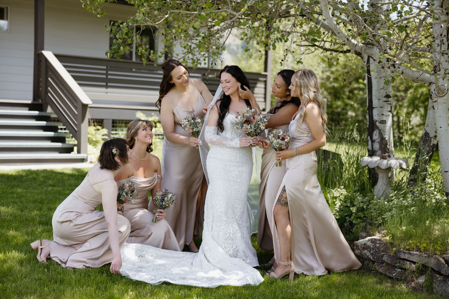 Bride in lace wedding gown surrounded by bridesmaids in champagne satin dresses holding bouquets outdoors.