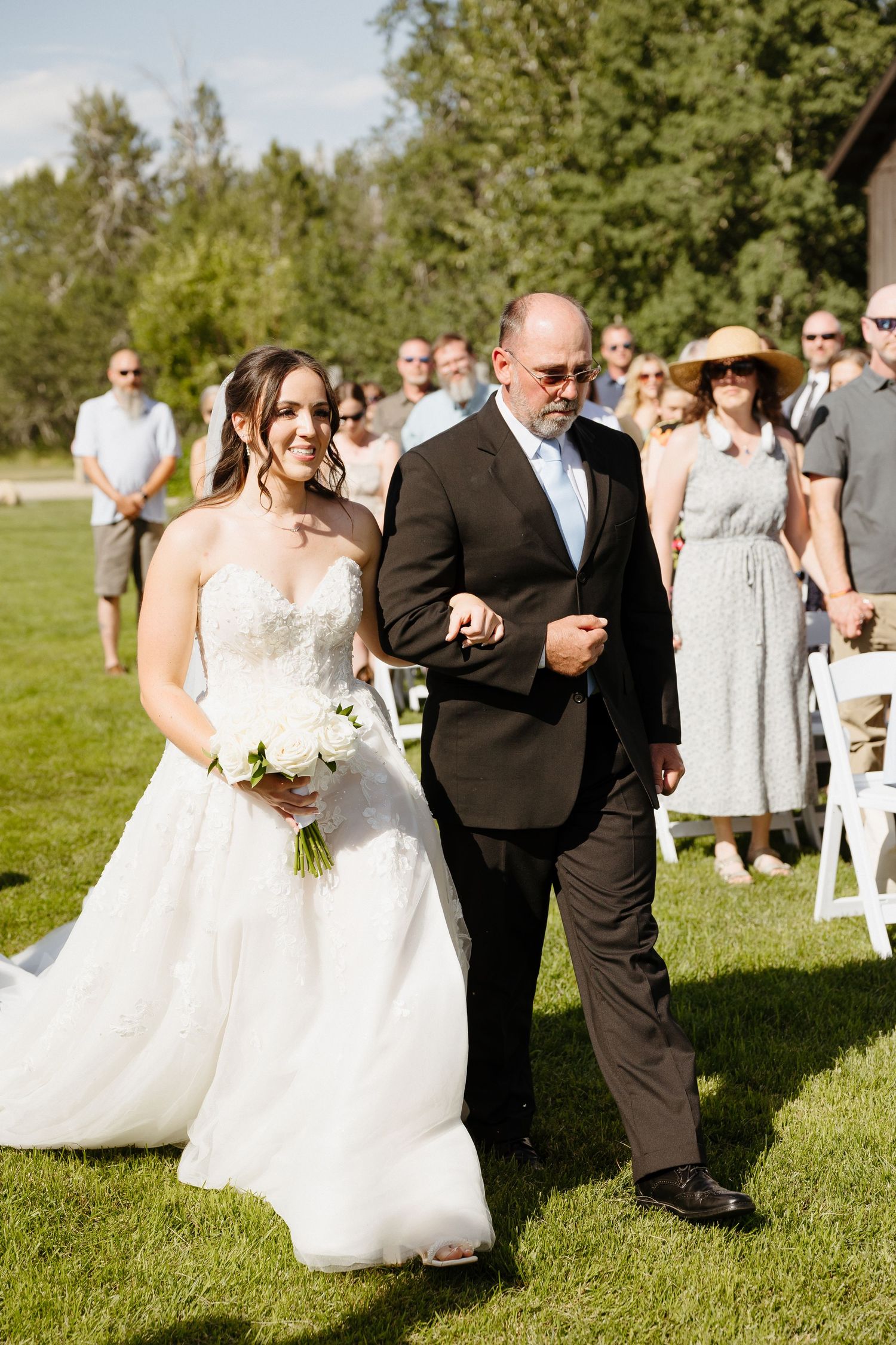 Bride in strapless lace gown walks down outdoor aisle with father at sunny wedding.