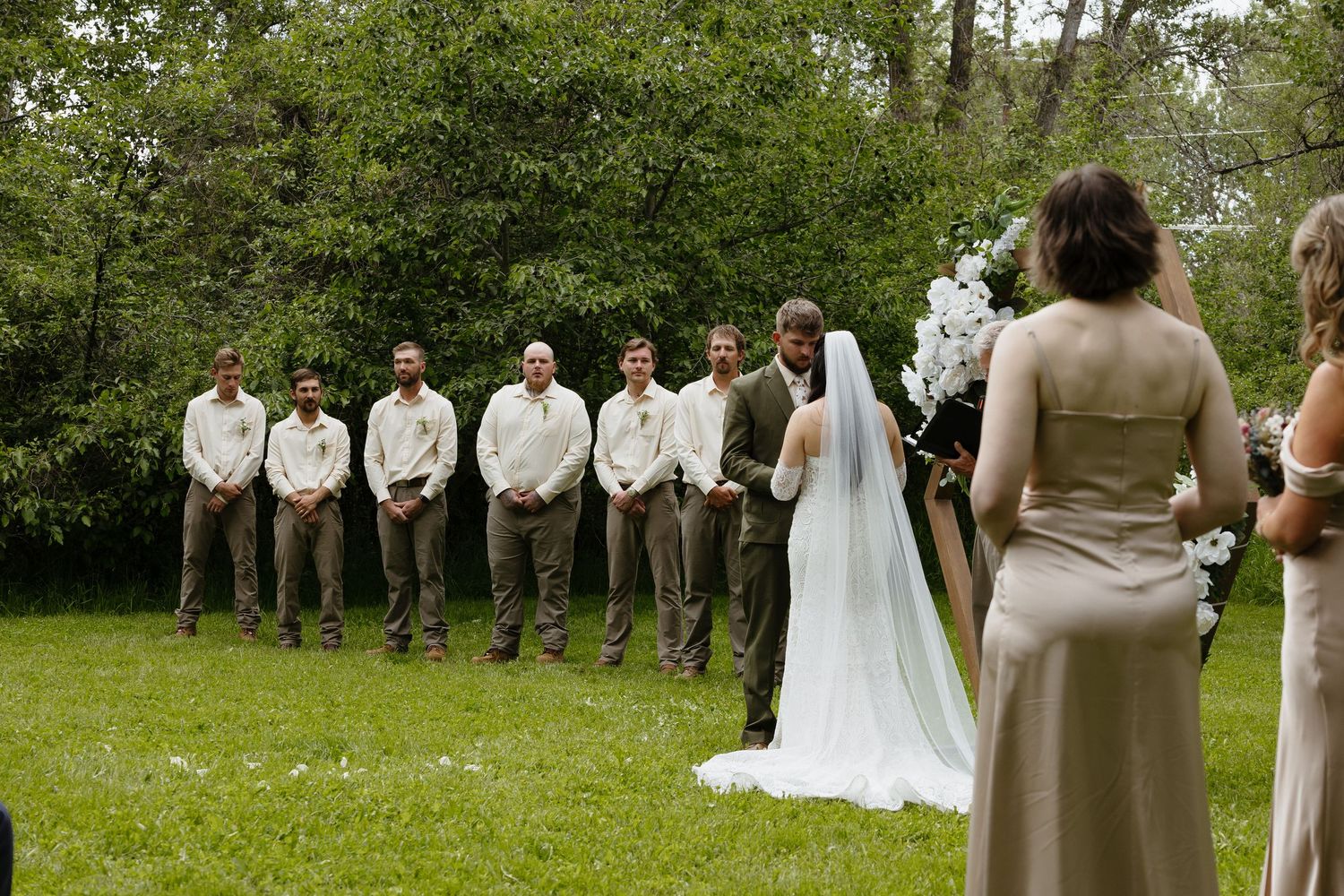 Groomsmen lineup in tan and olive suits at outdoor garden wedding ceremony.