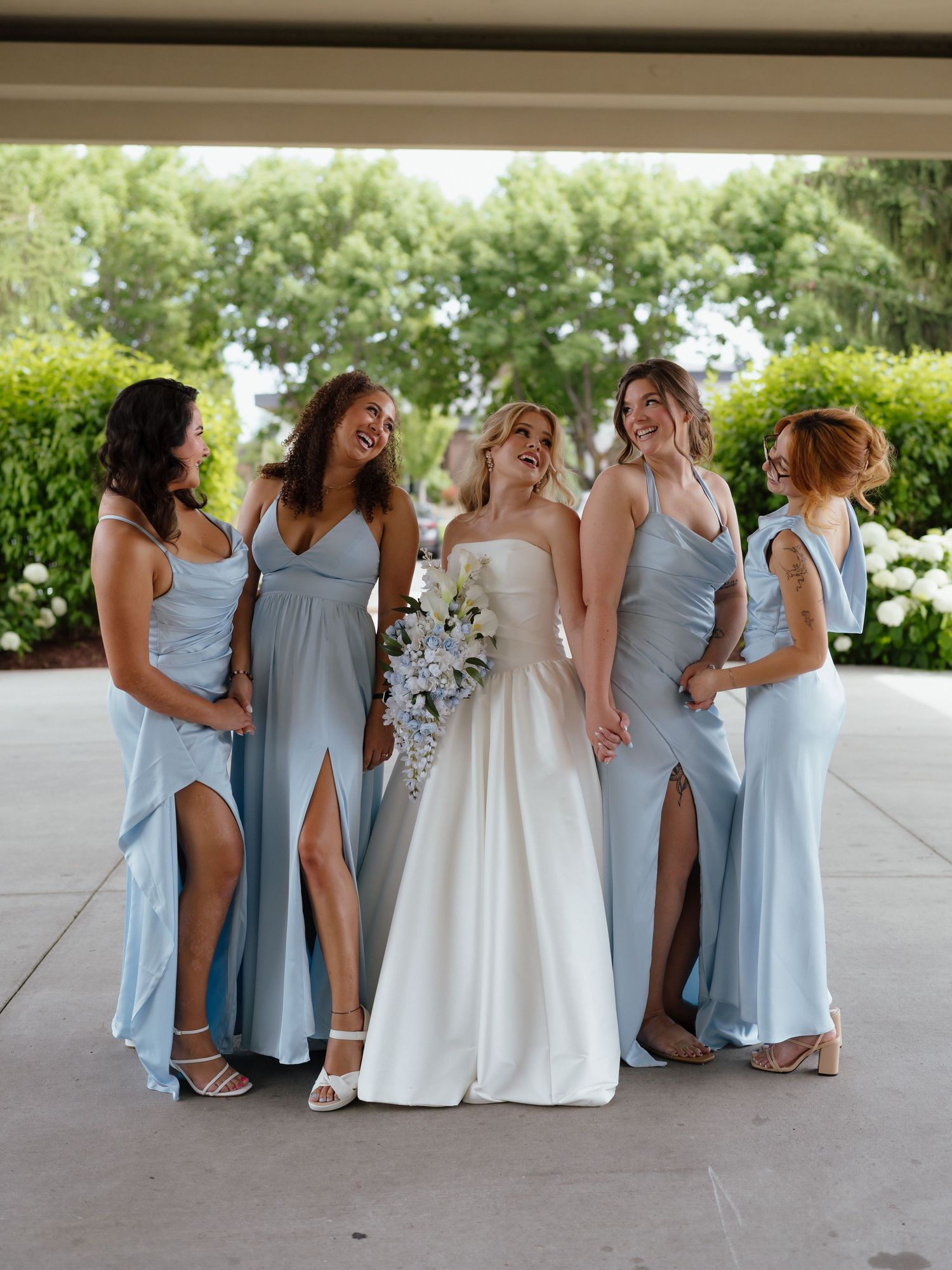 Bride in white strapless gown holding bouquet laughing with four bridesmaids in light blue dresses outdoors.