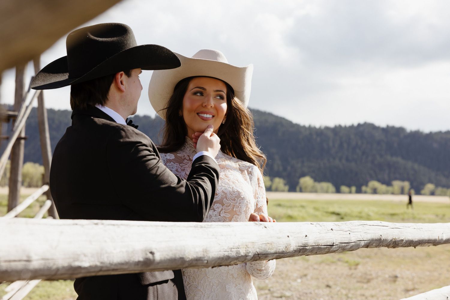 Couple in western cowboy hats sharing a romantic moment by a ranch fence with mountain views.