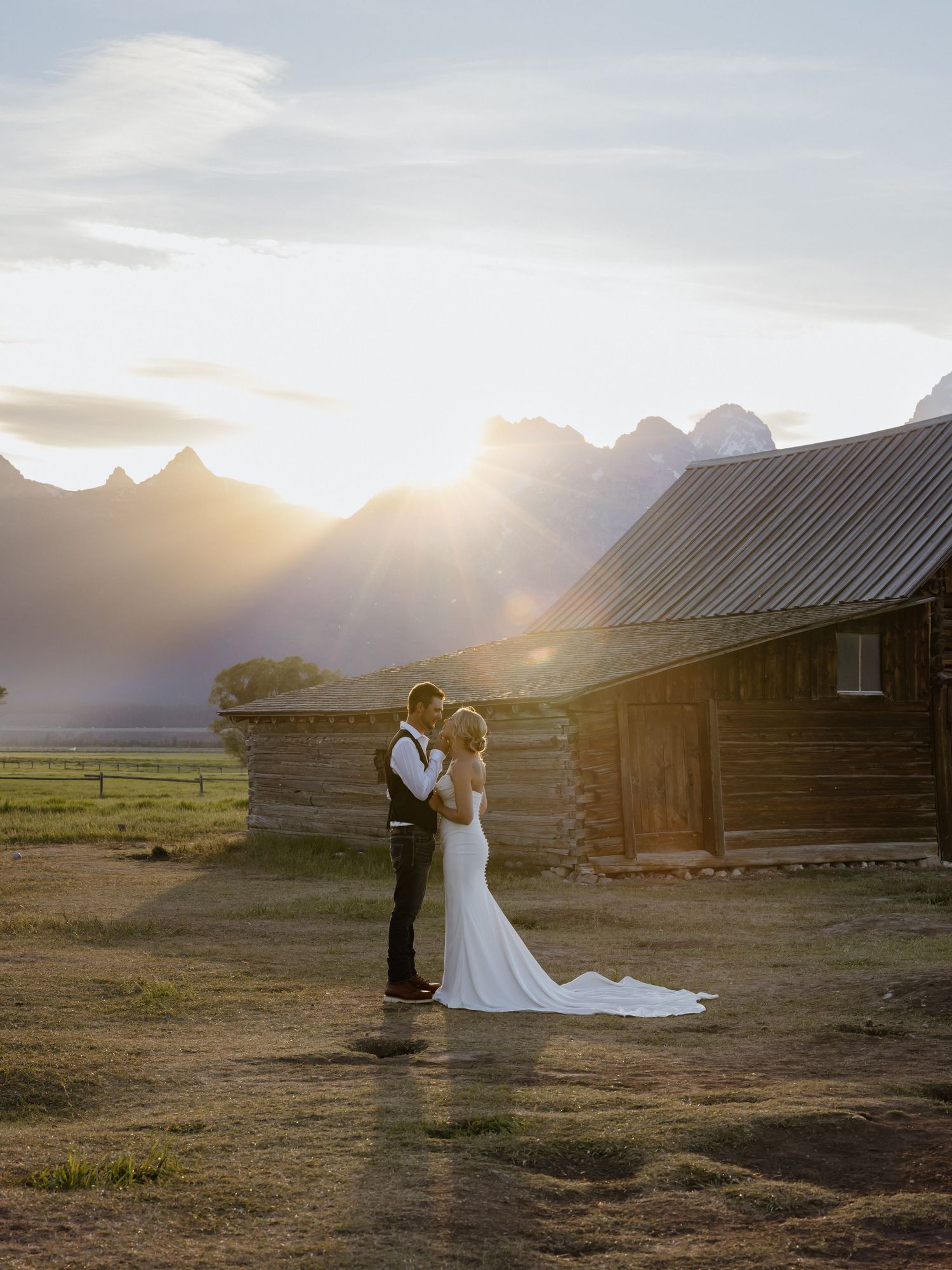 Bride and groom embrace at golden sunset near rustic log cabin with Grand Teton mountains backdrop.