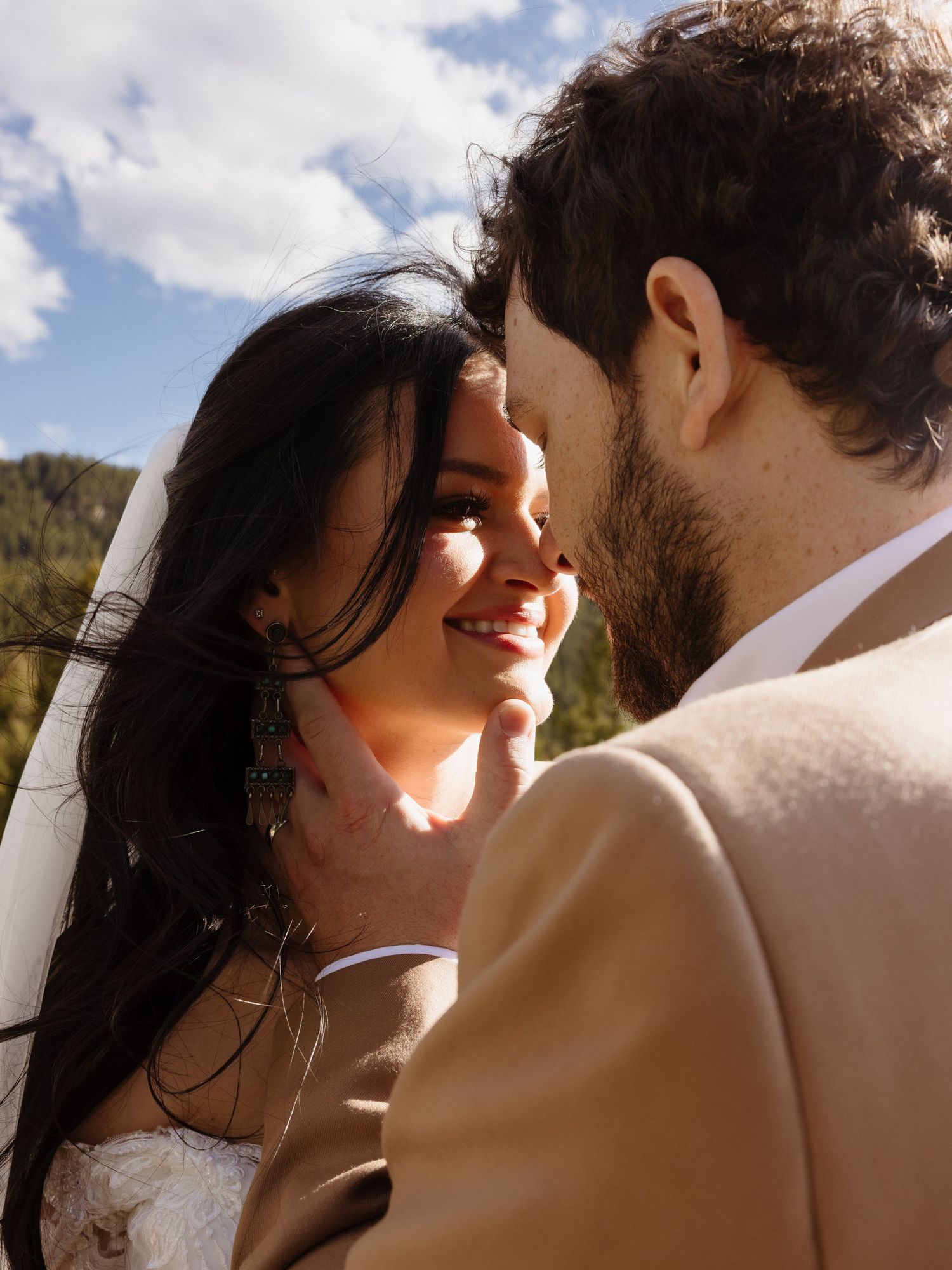 Groom tenderly cups brides face as she smiles in outdoor mountain wedding portrait.