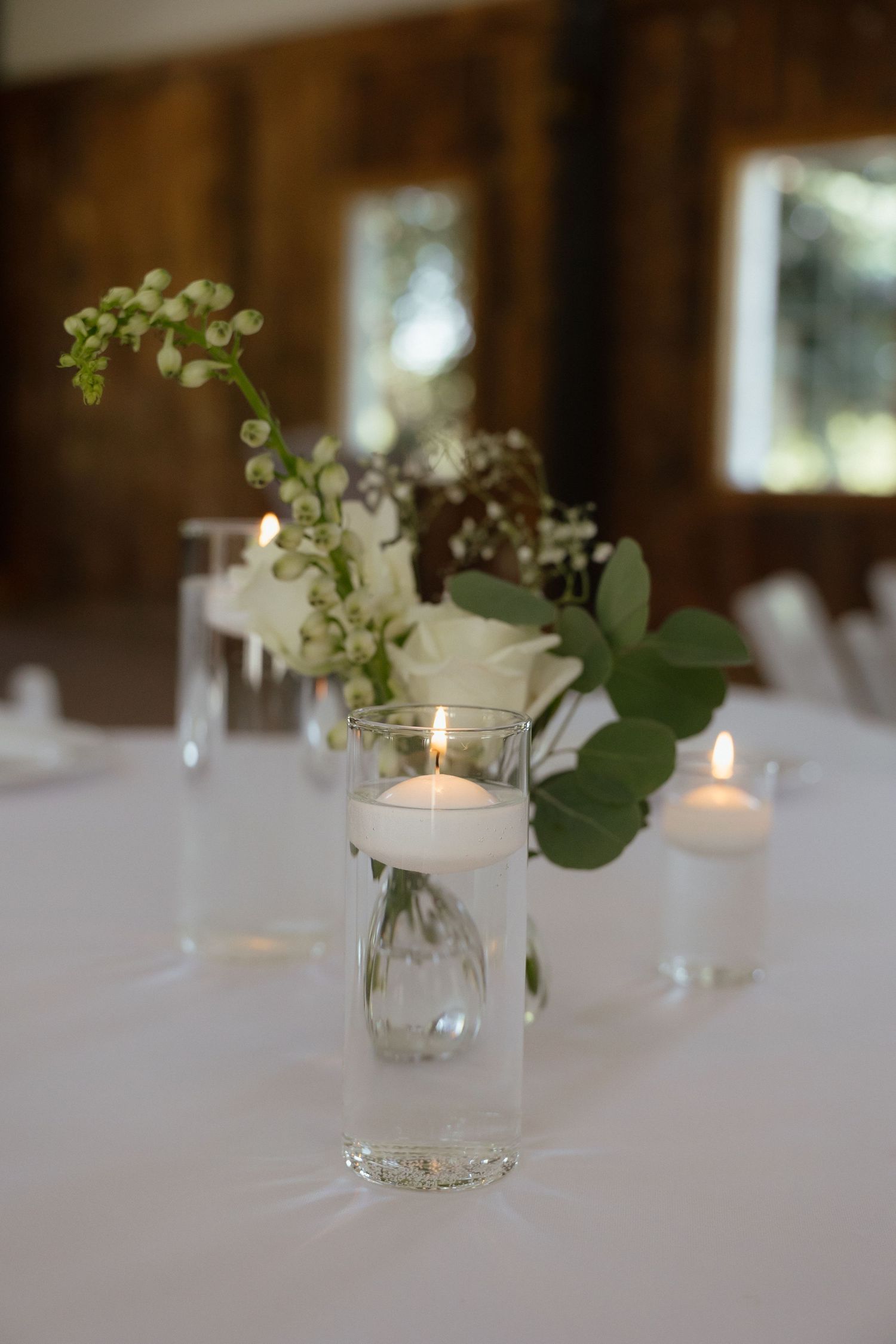 Elegant wedding table centerpiece with floating candle, white roses, and greenery in glass vases.