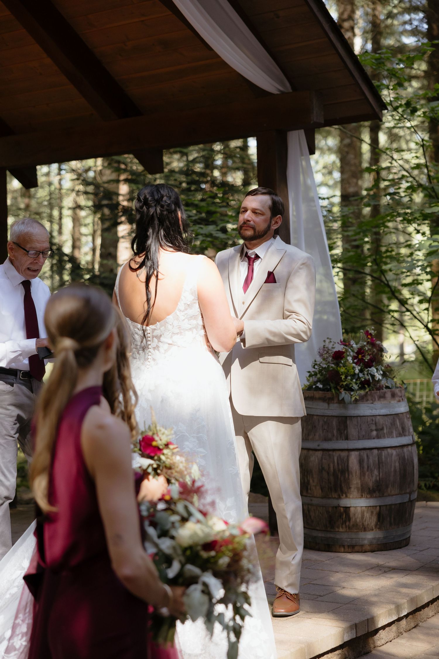 Groom in tan suit holds brides hands during outdoor forest wedding ceremony beneath a wooden pergola with barrel decor.