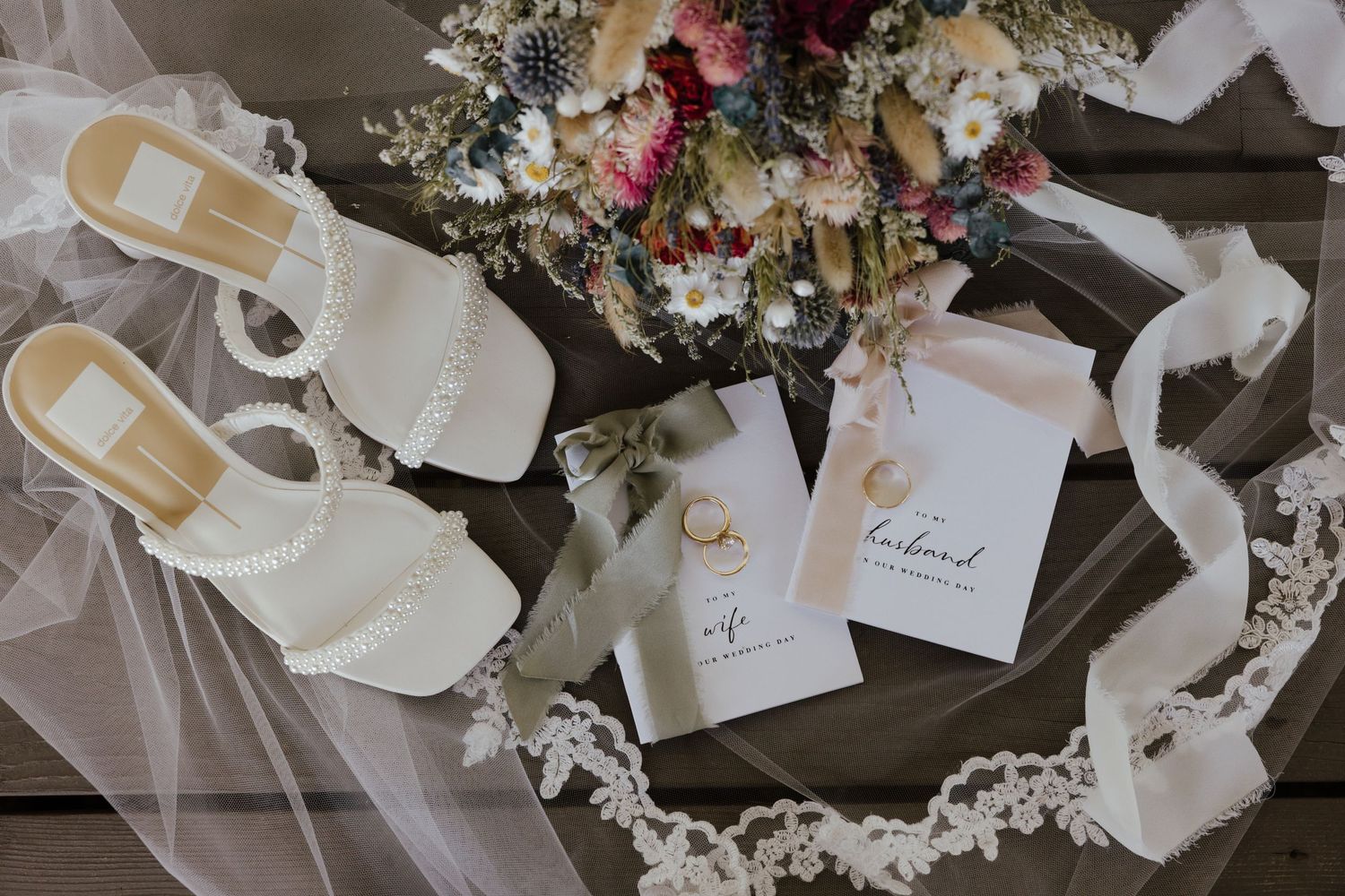 Elegant wedding flat lay with pearl heels, bridal veil, wildflower bouquet, and husband and wife vow books on rustic wood.