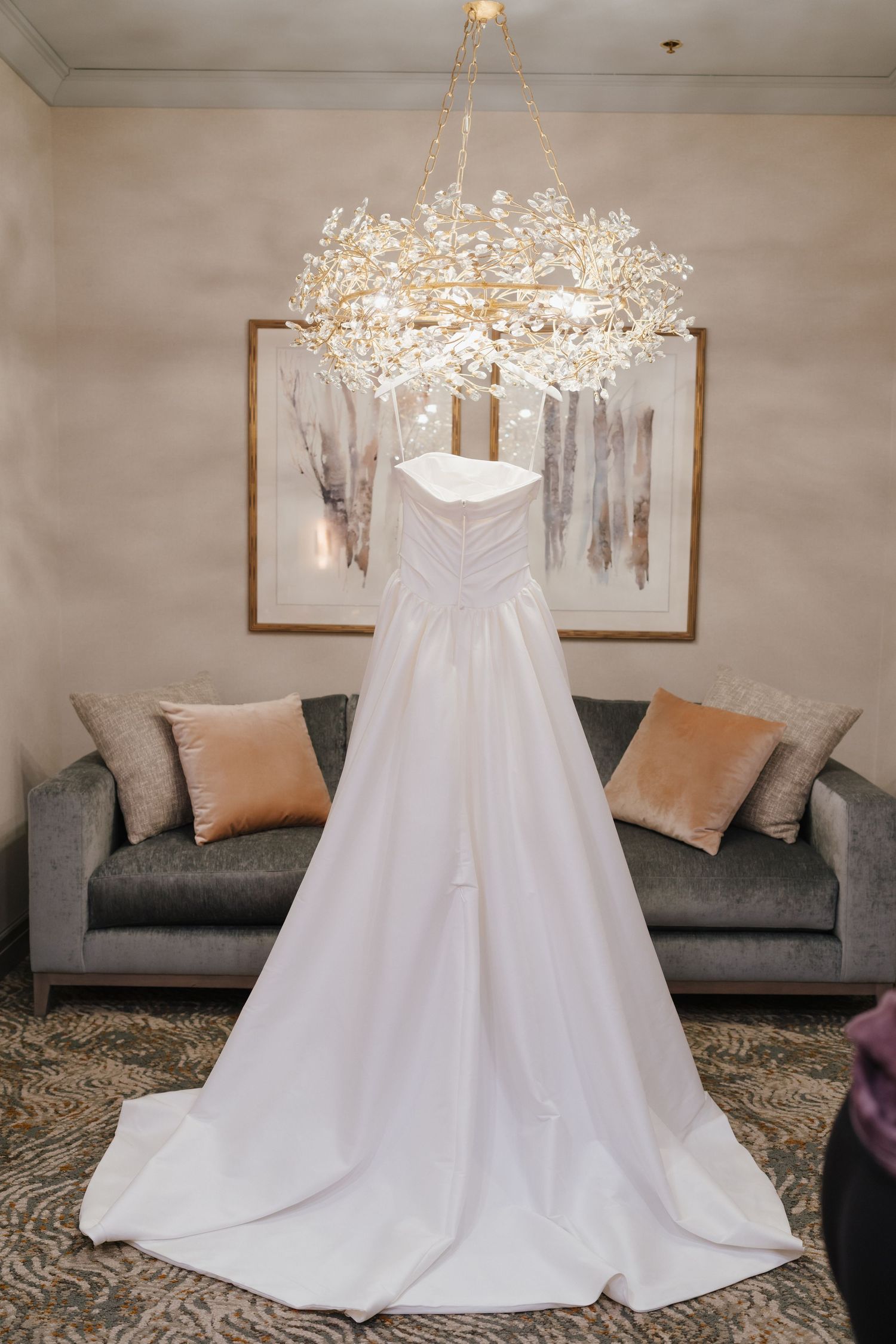 White wedding gown with long train displayed on a hanger beneath an elegant crystal chandelier in a luxurious hotel suite.