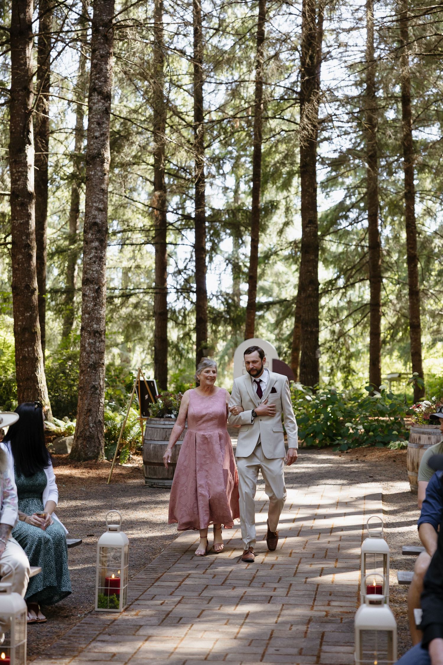 Groom walking his mother down the brick aisle at an outdoor forest wedding ceremony surrounded by tall pine trees.