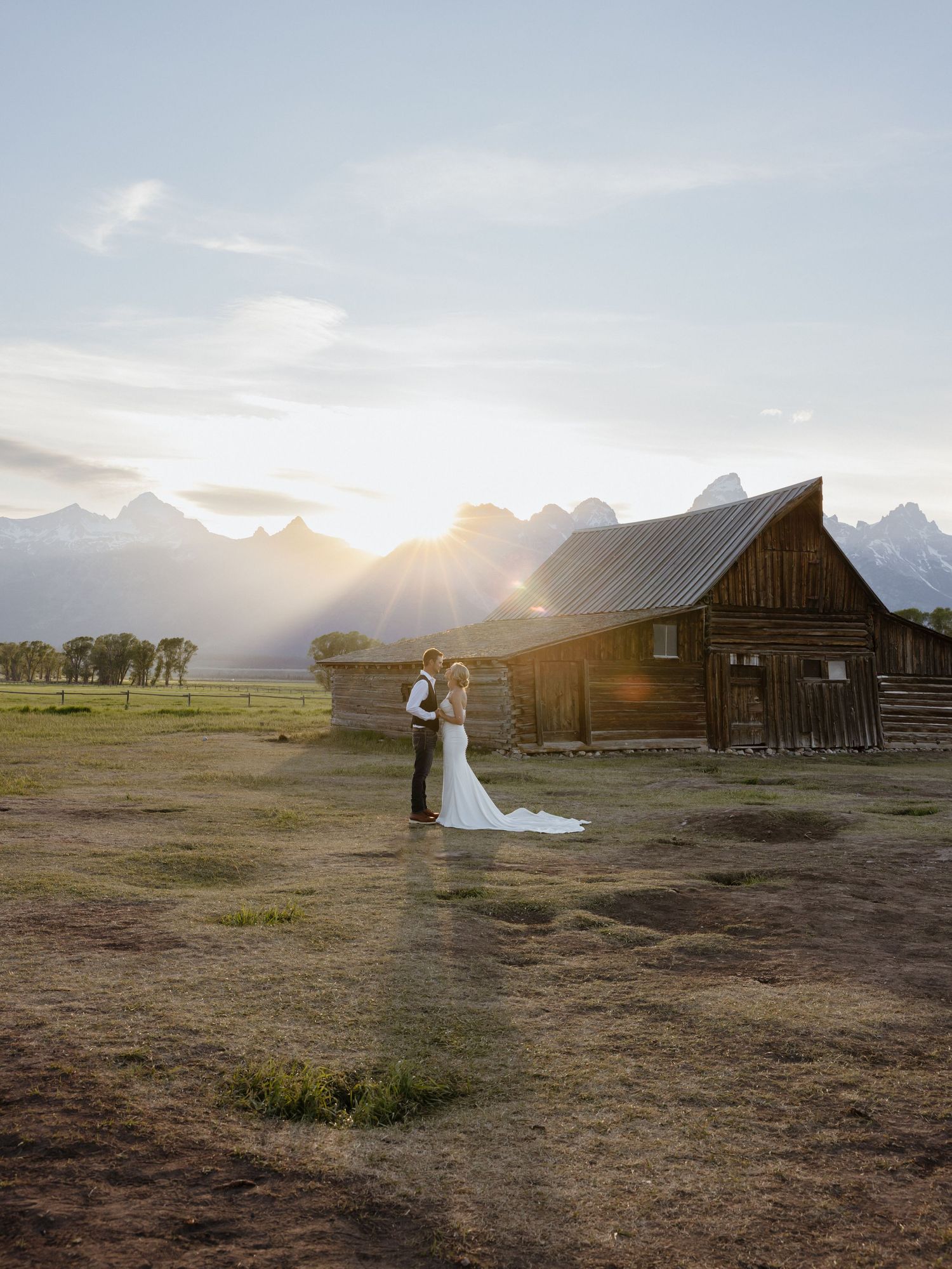 Bride and groom embrace near rustic Mormon Row barn with Grand Teton mountains glowing at sunset.