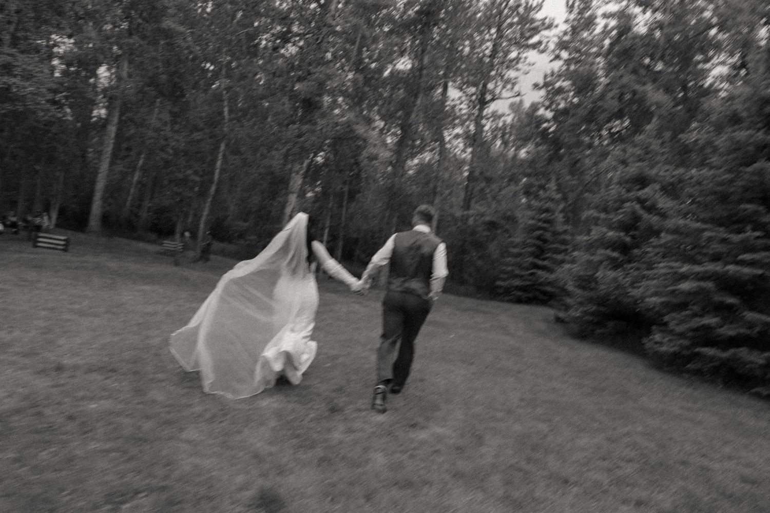 Black and white photo of bride and groom running hand in hand through lush green forest.