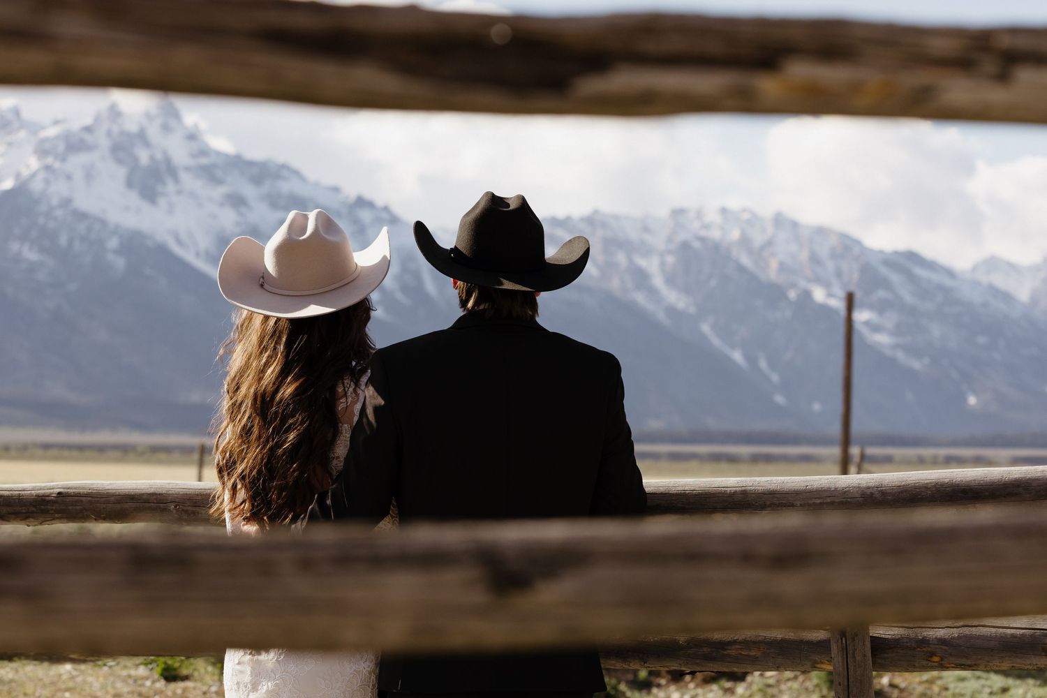 Bride and groom in cowboy hats standing together facing snow-capped mountains at a wooden fence.