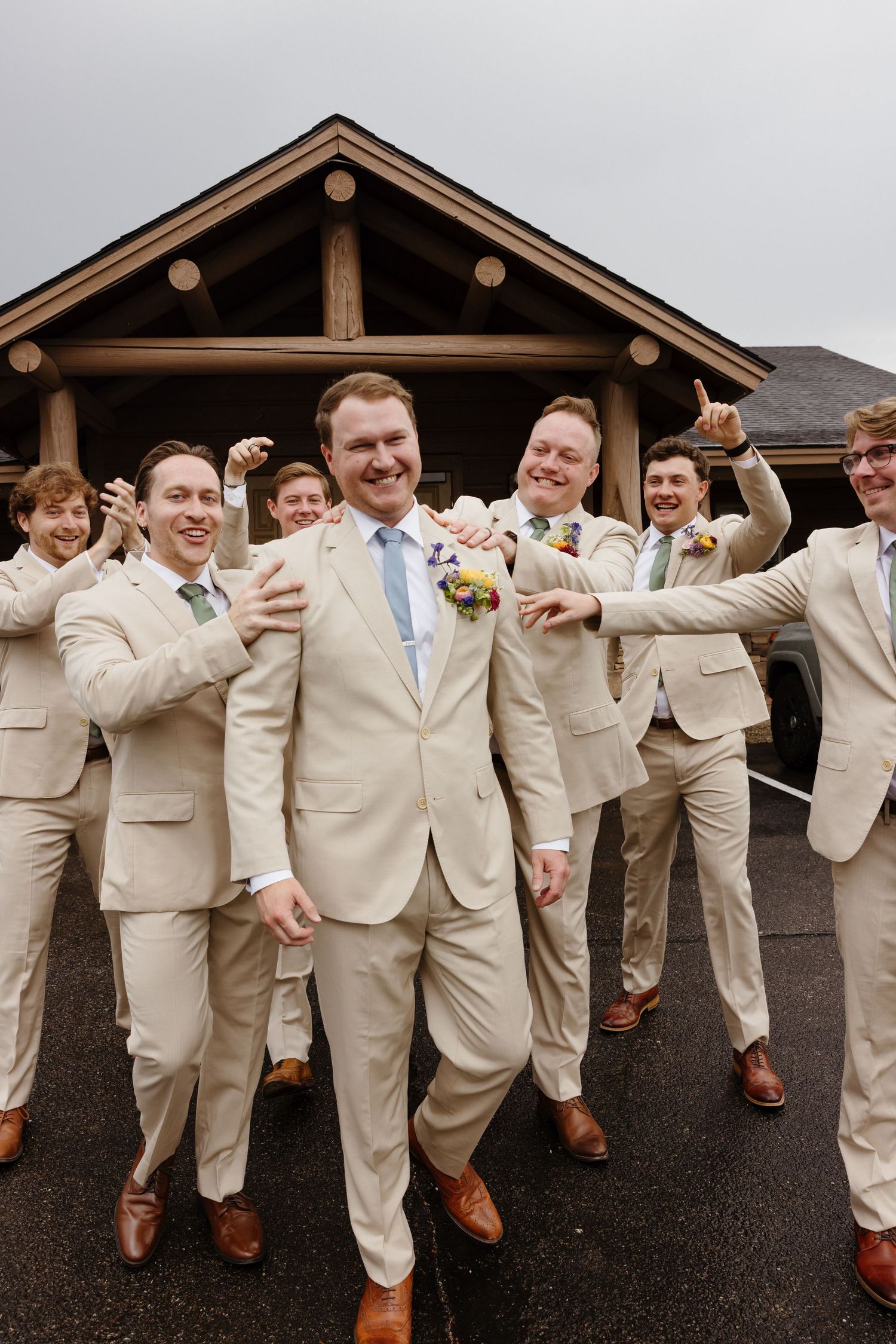 Groom in a tan suit laughs as his groomsmen in matching suits celebrate together outside a rustic lodge venue.