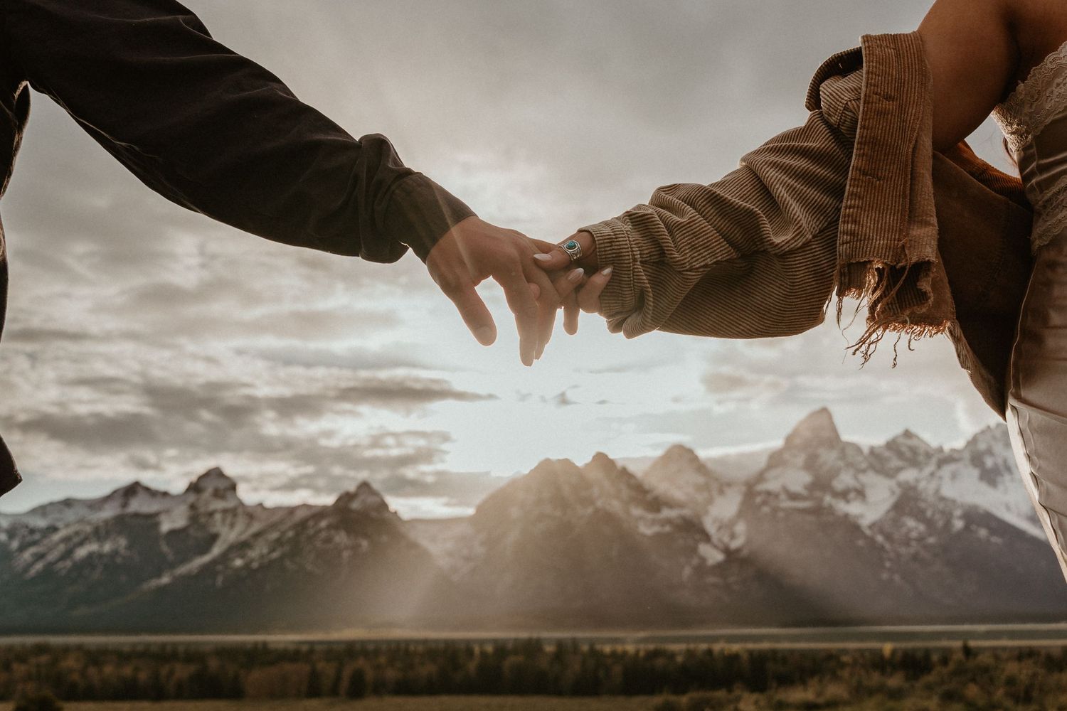 Close-up of couple holding hands with fingers touching against mountain landscape at sunset.