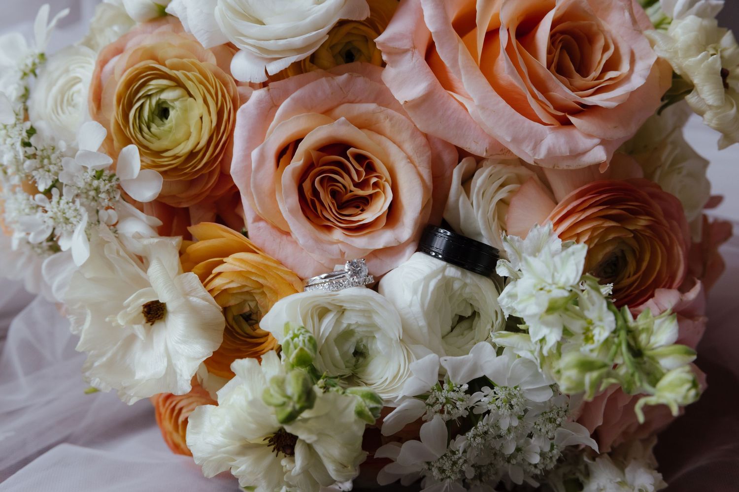 Close-up of bridal bouquet with peach roses and white ranunculus with wedding rings nestled inside.