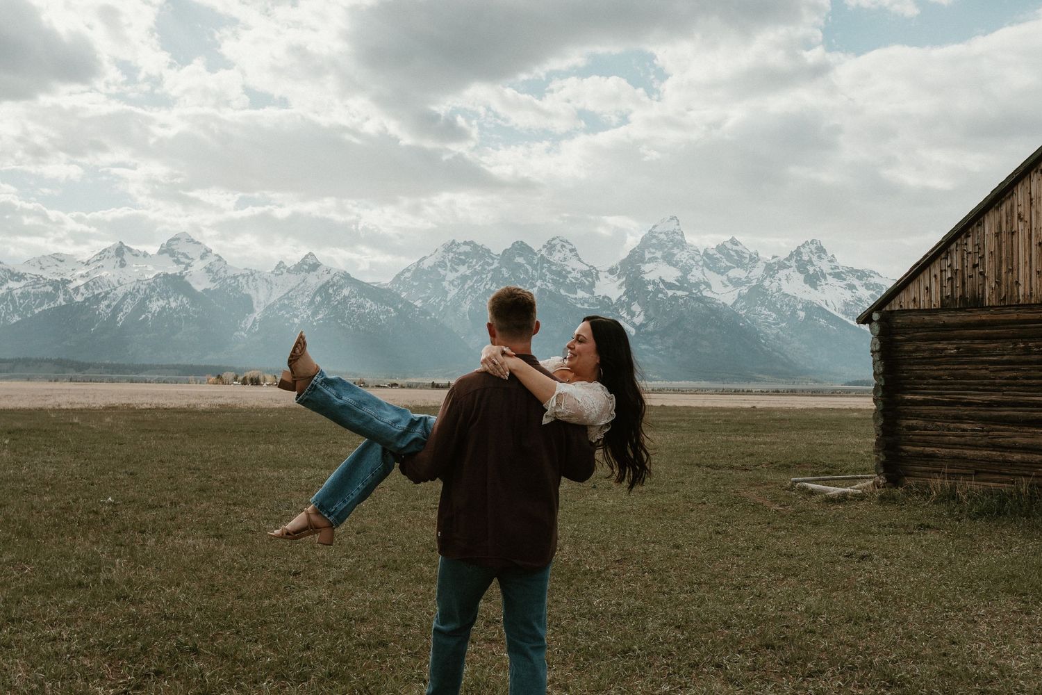 Man carrying woman in his arms in a field with a historic barn and Grand Teton mountains behind them.