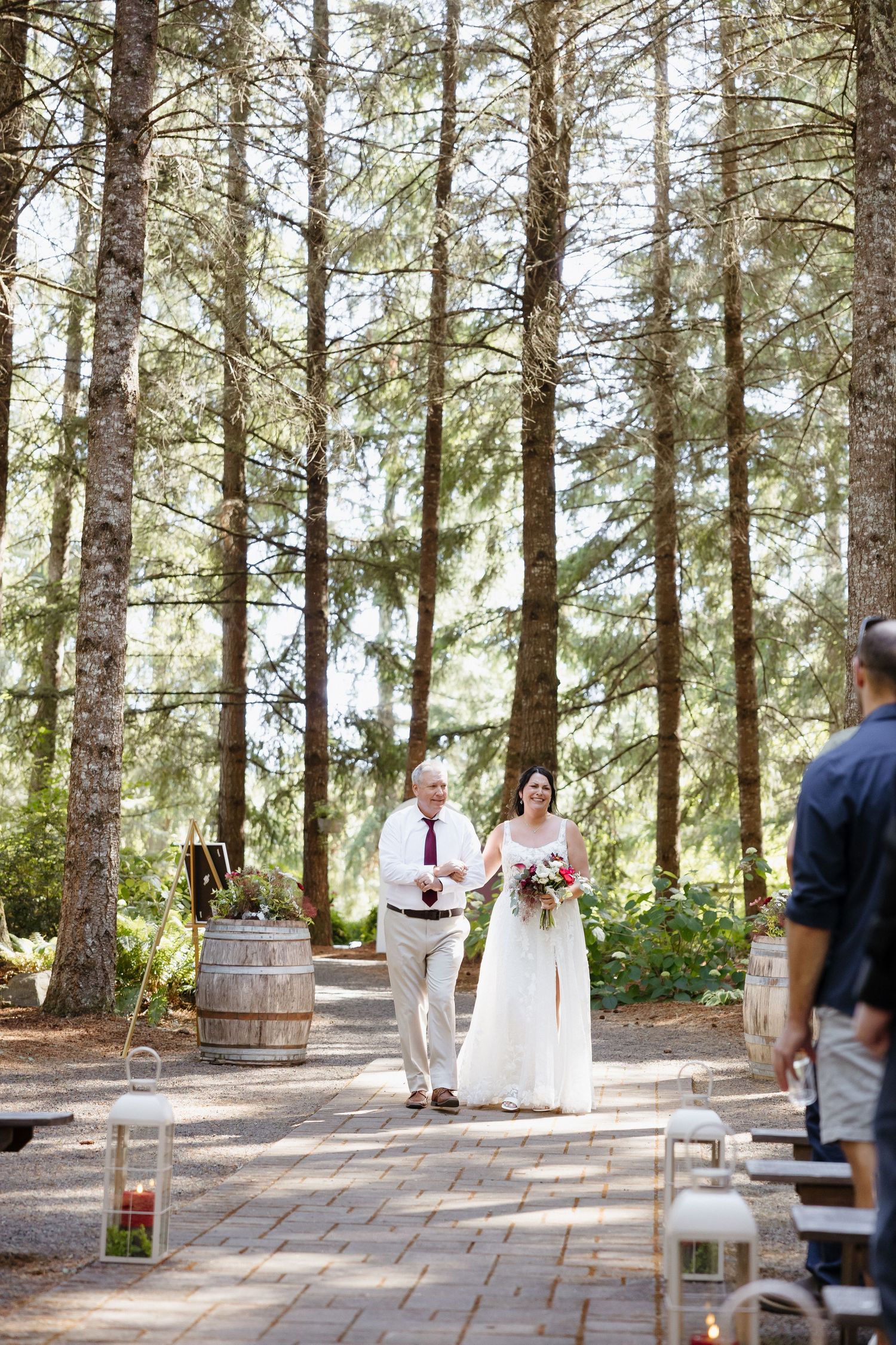 Father walking bride down a sunlit forest aisle with tall pine trees and lanterns lining the path.