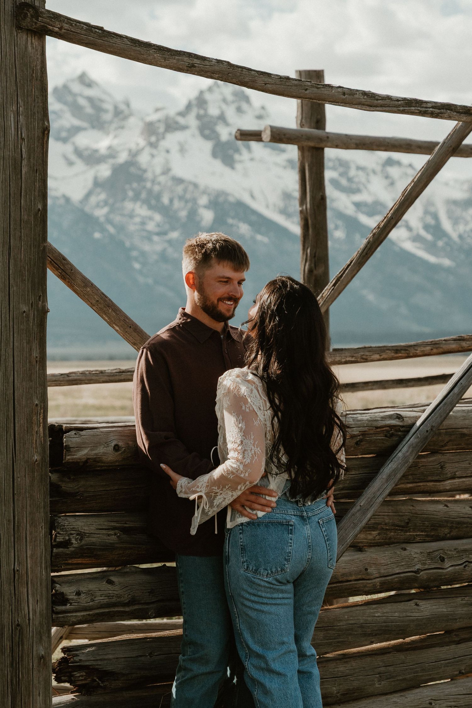 Couple sharing a tender moment under a wooden structure with snow-capped mountains visible in the background.