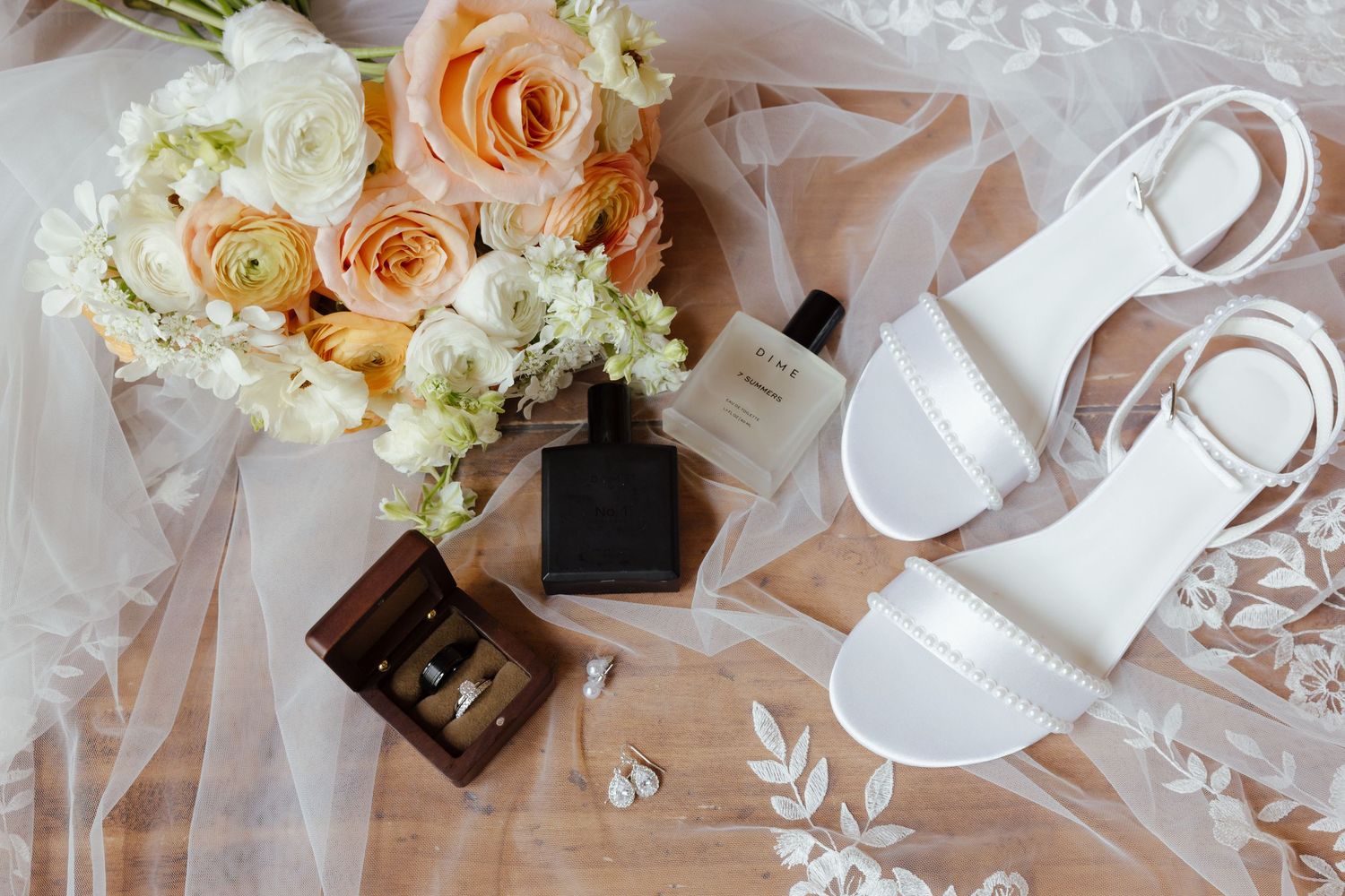 Bridal flat lay featuring white sandals, peach roses bouquet, rings, veil, and wedding accessories.