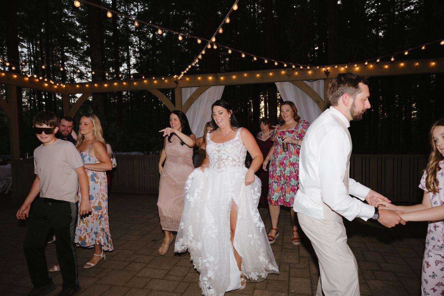 Bride in lace wedding dress dancing joyfully with guests under string lights at outdoor evening wedding reception.