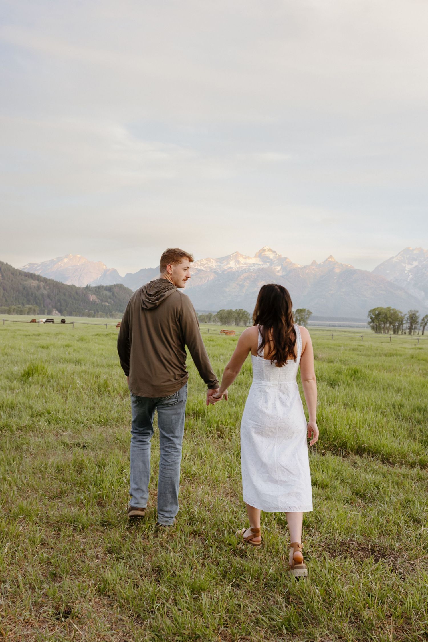 Couple holding hands walking through a lush green meadow toward the Grand Teton mountains.
