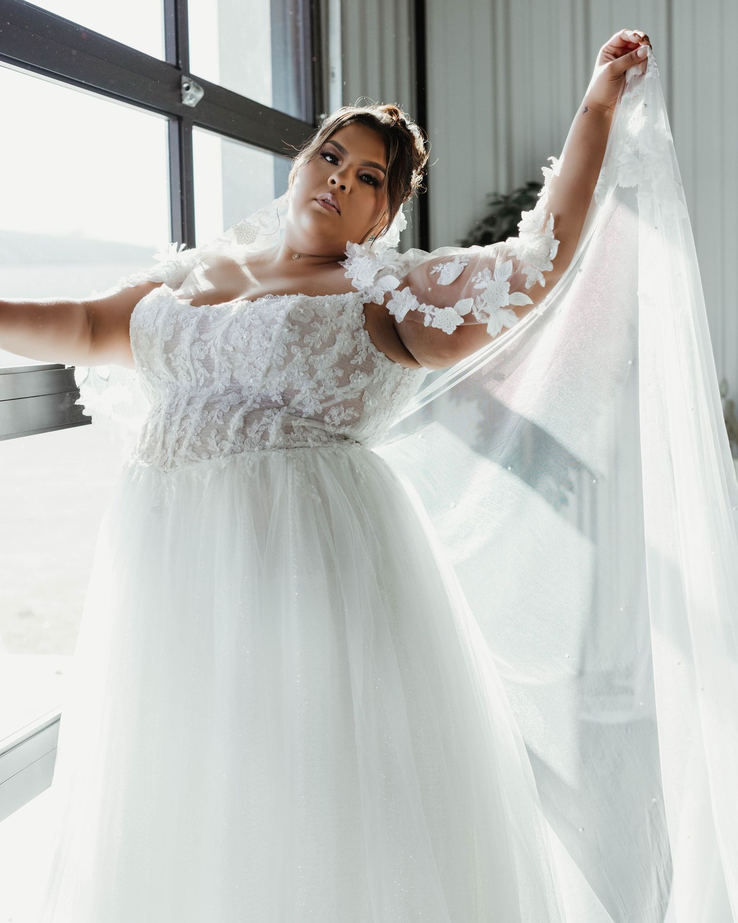 Plus-size bride in floral lace wedding gown holds flowing veil wide open near large venue windows.