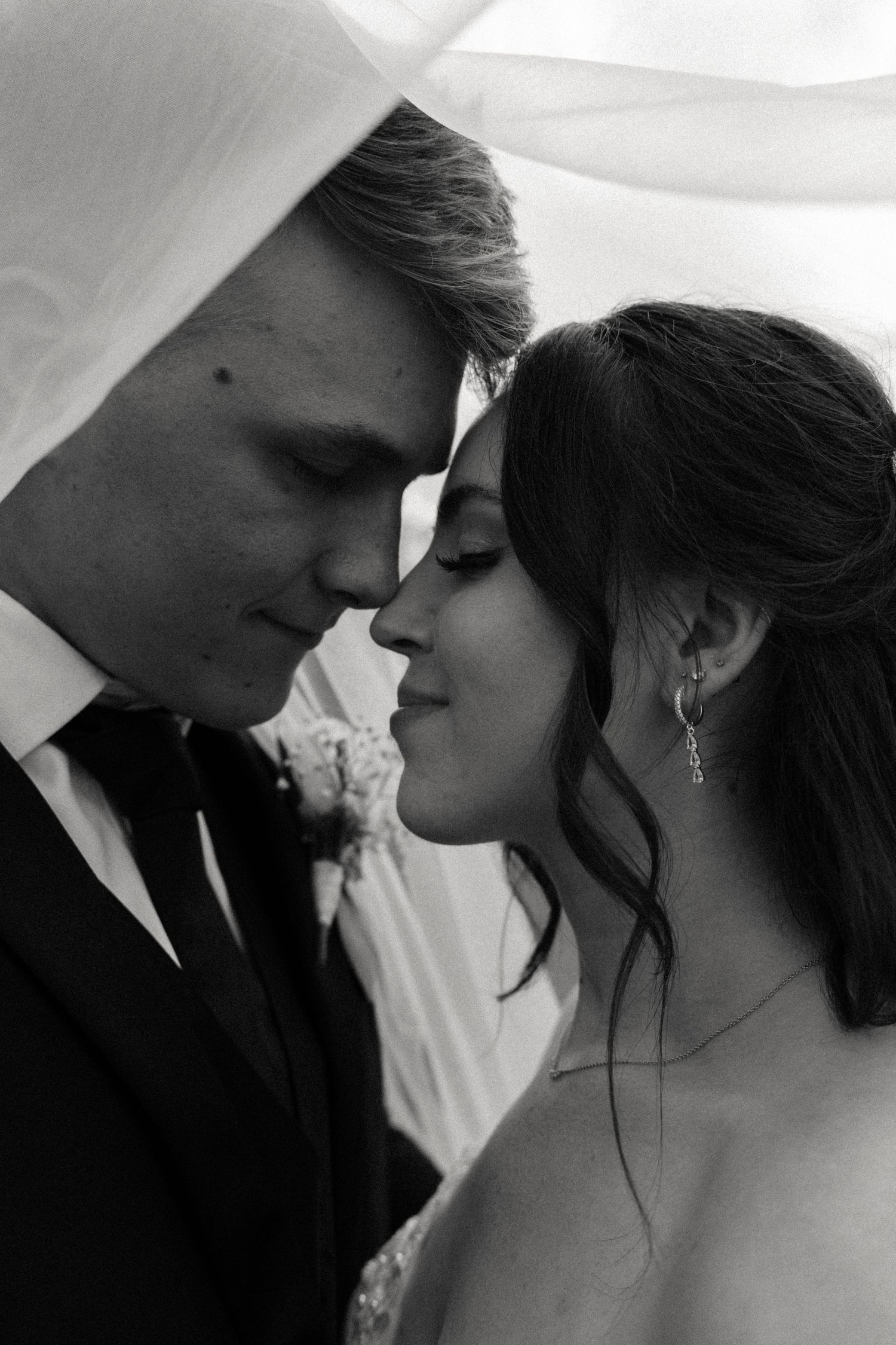 Intimate black and white portrait of a bride and groom touching foreheads tenderly beneath a flowing wedding veil.