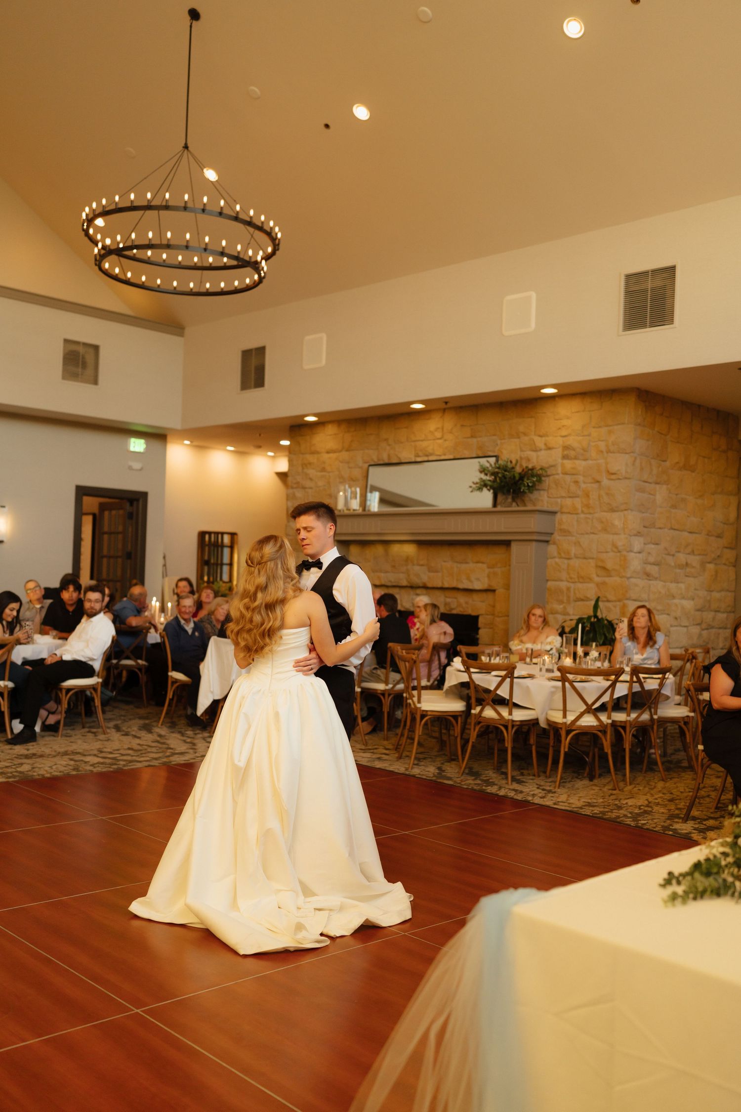 Bride and groom share their first dance at elegant wedding reception with stone fireplace and chandelier.