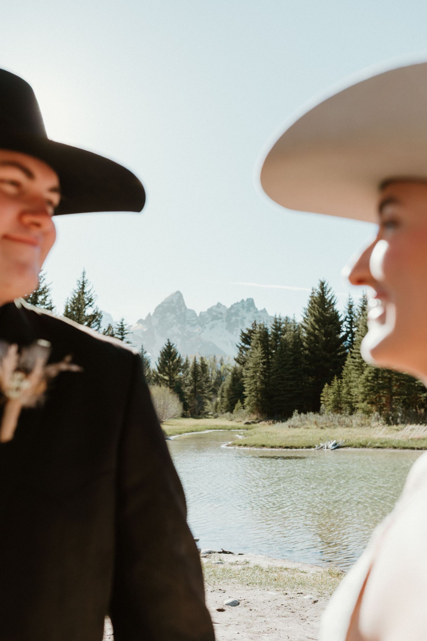 Smiling groom in black cowboy hat faces bride in white hat with Grand Teton mountains visible between them.