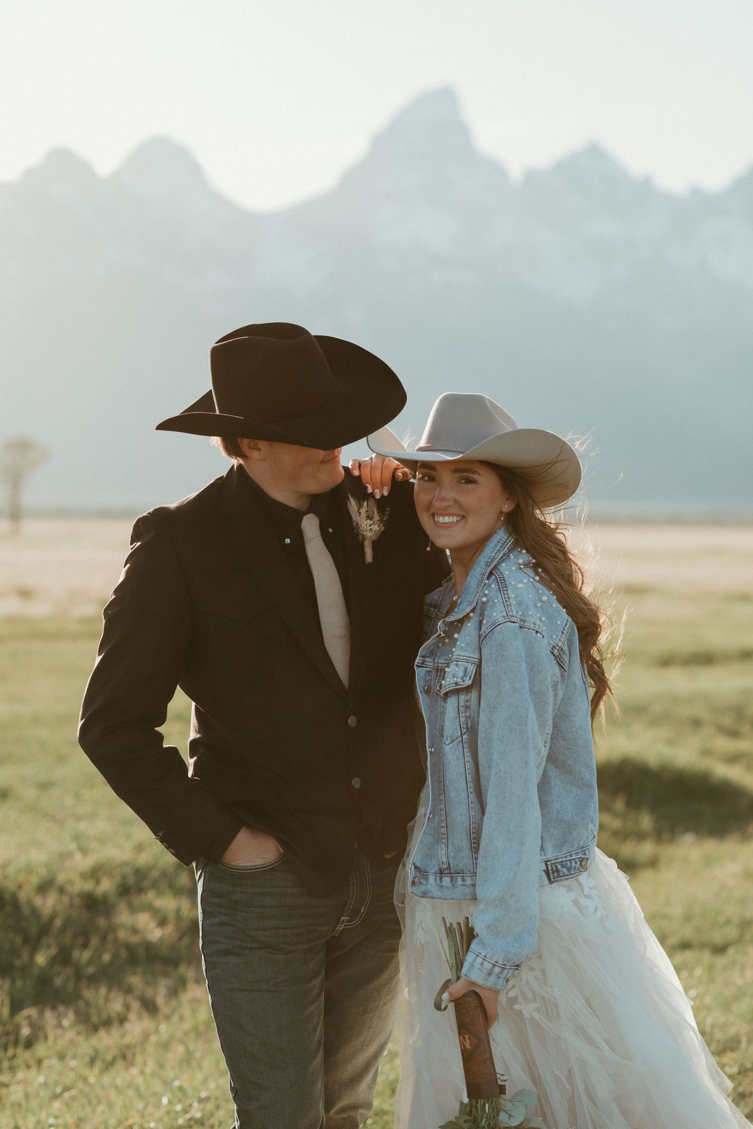 Smiling bride in denim jacket and white cowboy hat leans on groom in black hat at Tetons.