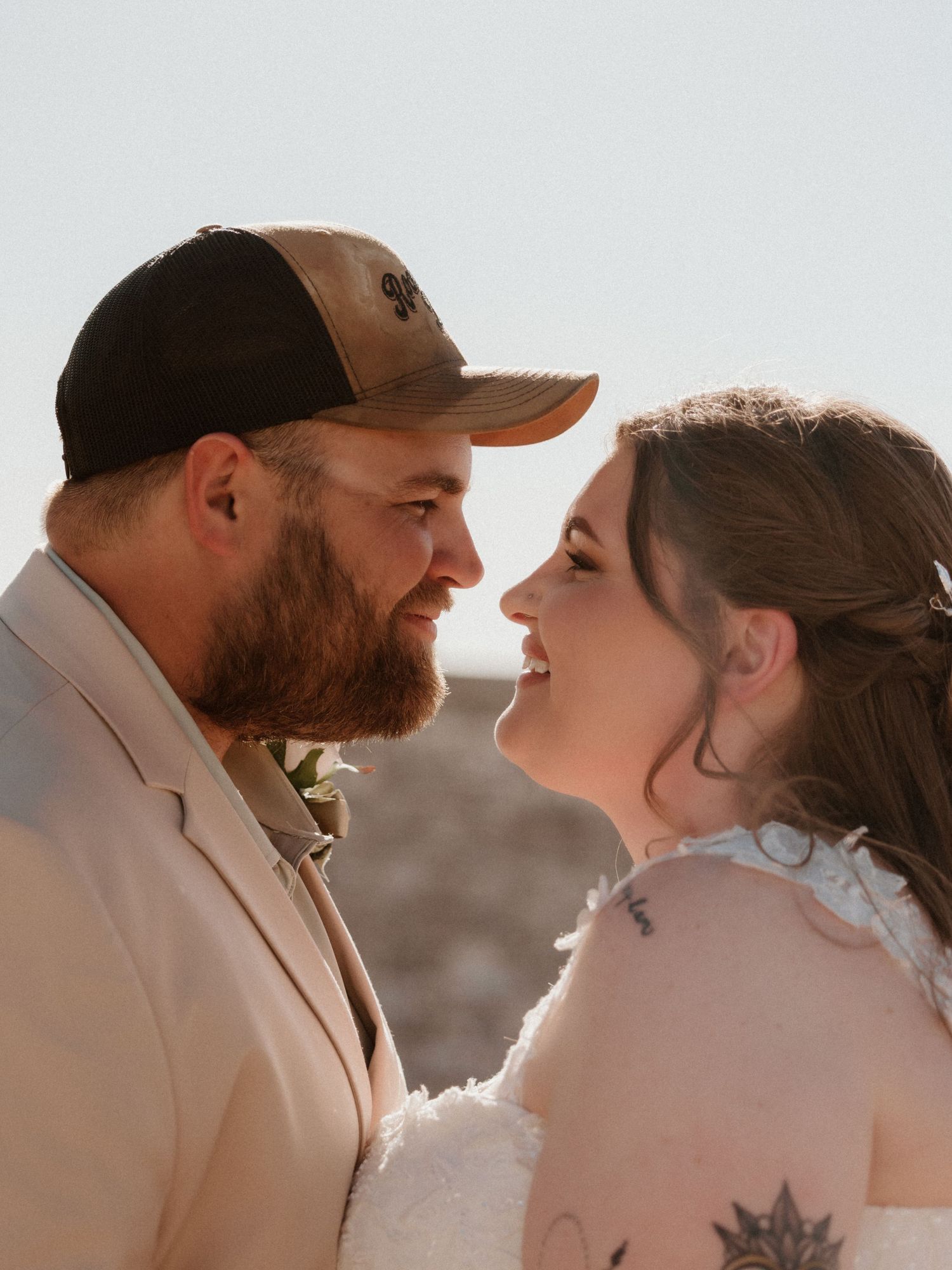 Smiling bride and bearded groom in baseball cap share an intimate moment at outdoor wedding ceremony.
