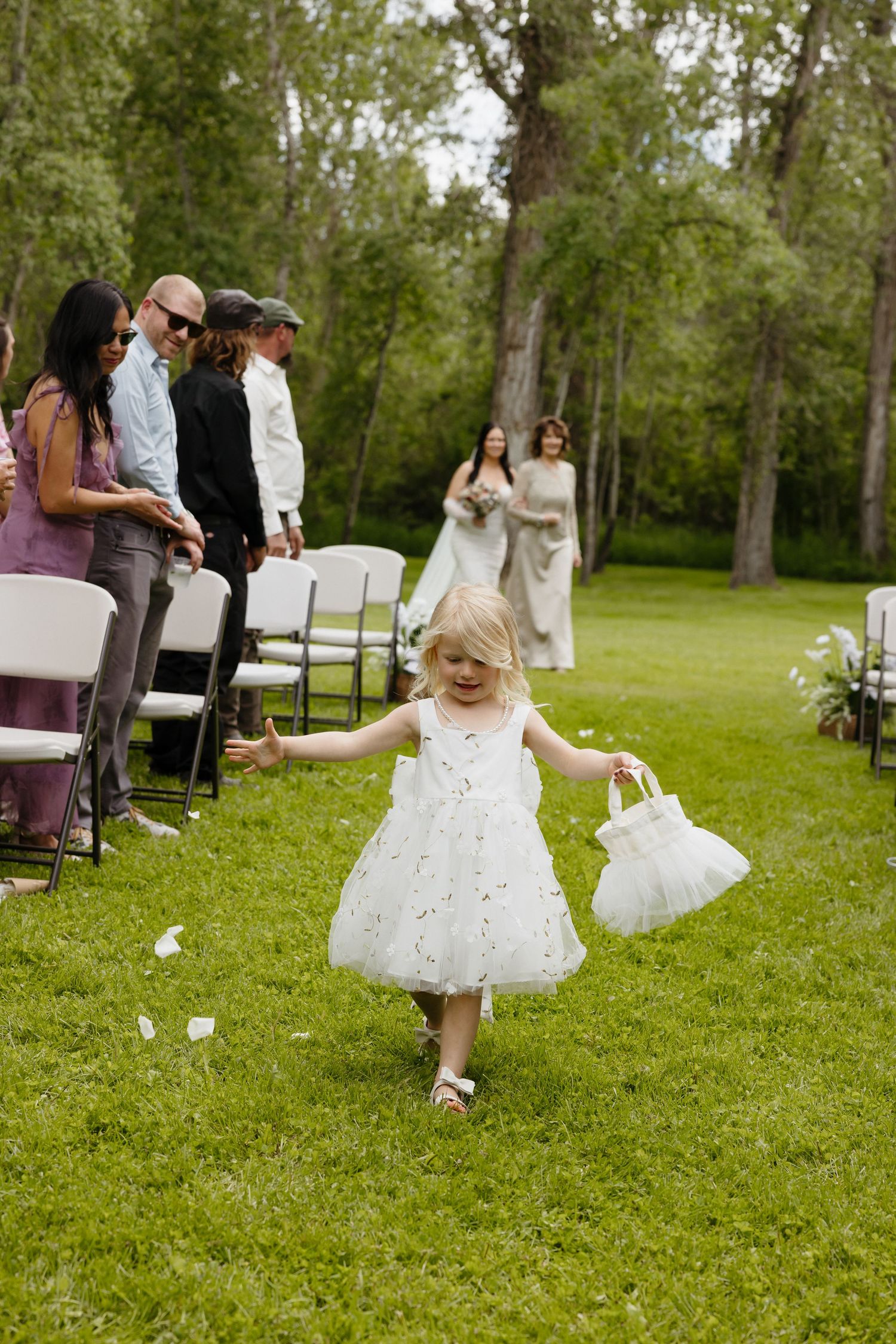 A flower girl in a white dress scatters petals down the aisle at an outdoor wedding ceremony.